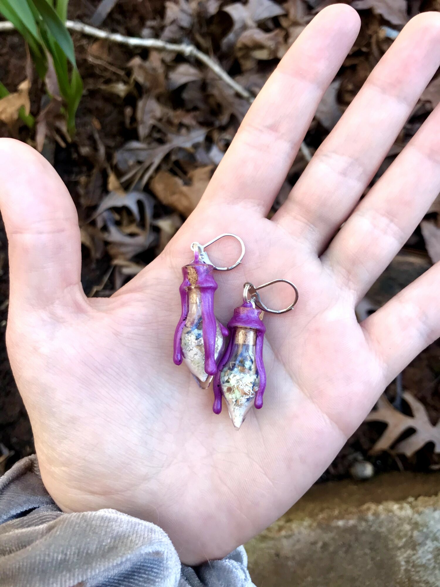 Hand holding two purple and white beaded earrings with purple drips, outdoors with leaves on the ground