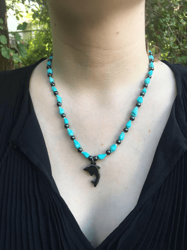 Person wearing a turquoise beaded necklace with a black pendant, black top, outdoors background with greenery