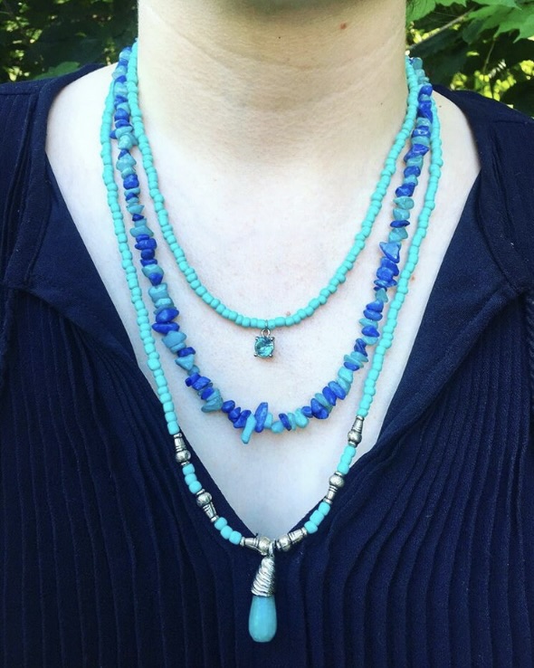 Person wearing multiple layered blue beaded necklaces over a dark top, outdoors with green foliage in background.