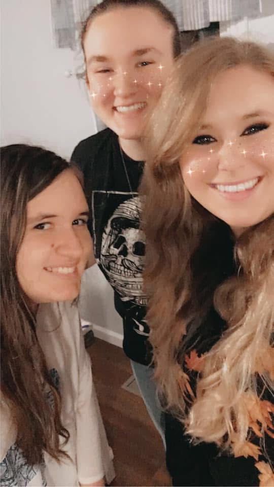 Three smiling women close together indoors, with light-colored walls and a window in the background.