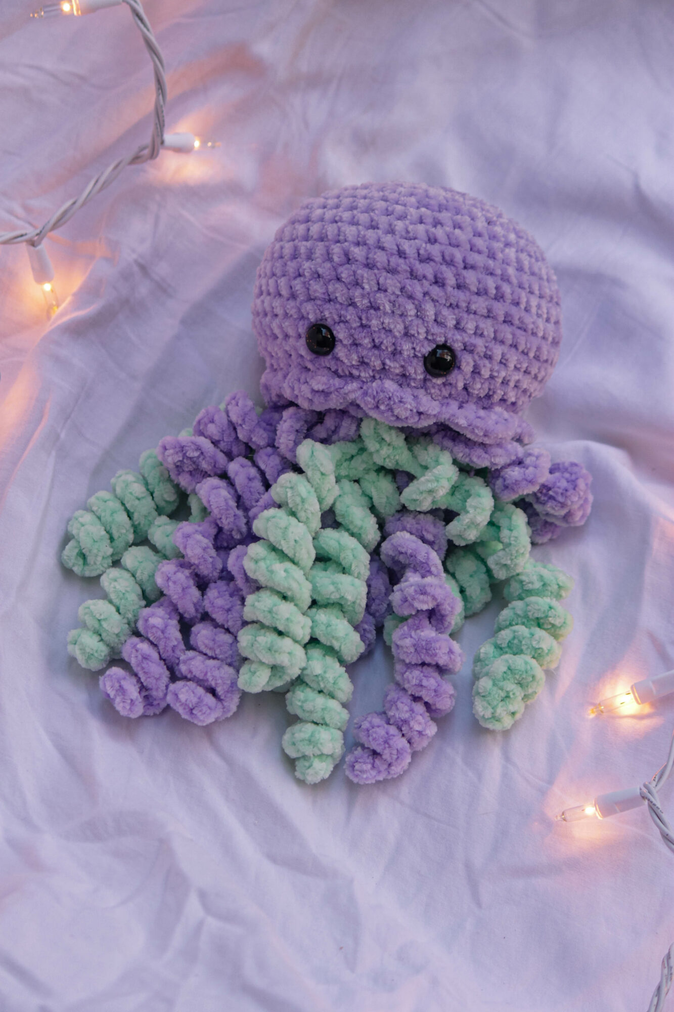 Purple and green crocheted octopus plush toy on white fabric with string lights, front view.