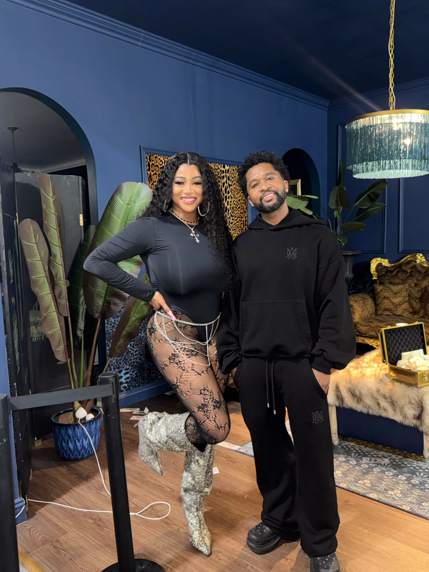 Two women pose indoors with blue walls, plants, and decorative items in the background. One woman wears a black dress with lace details, the other in black pants and a hoodie. Both are smiling.
