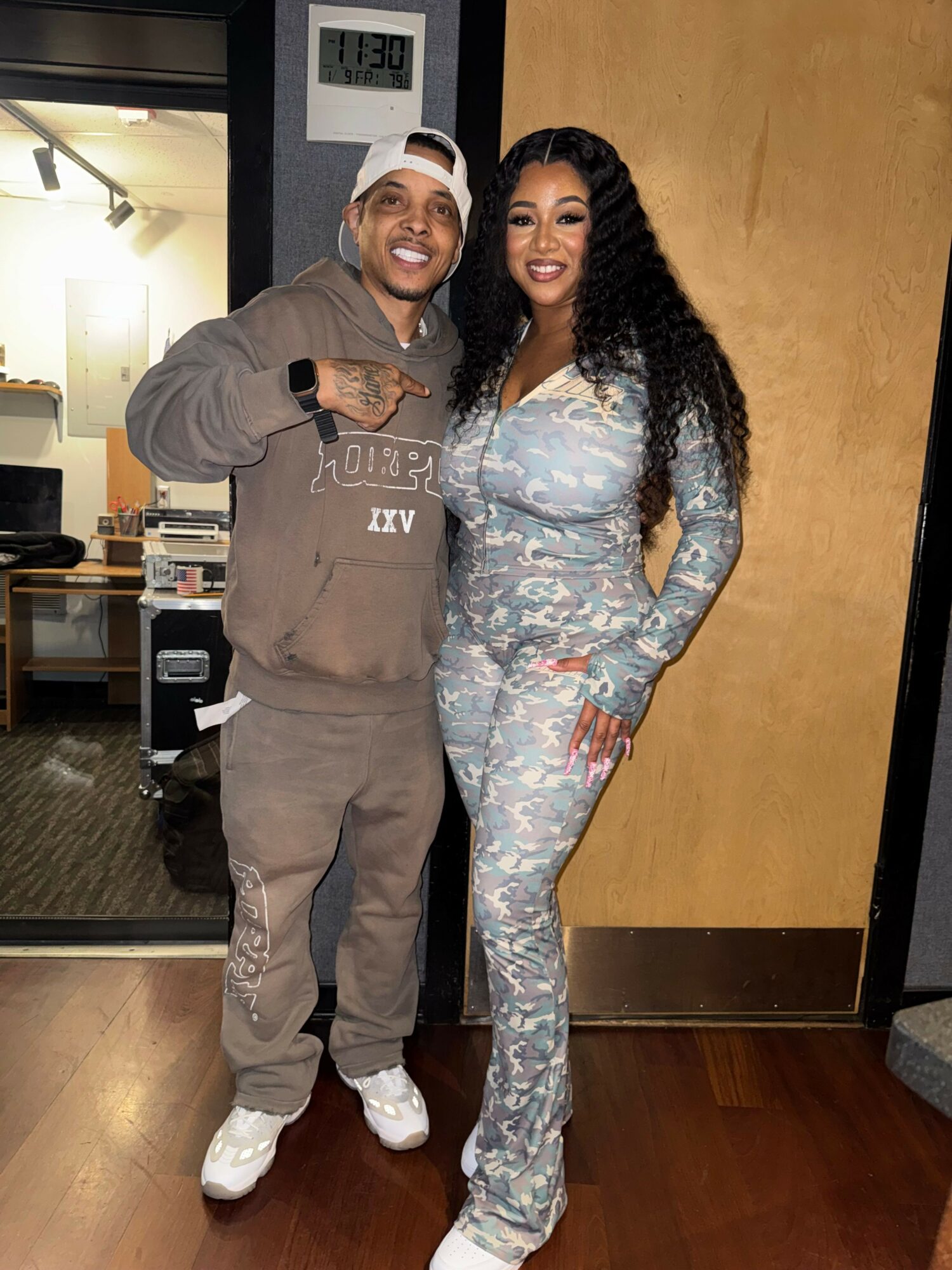 Two people standing indoors, one in a gray hoodie and the other in a patterned jumpsuit, smiling and taking a selfie.