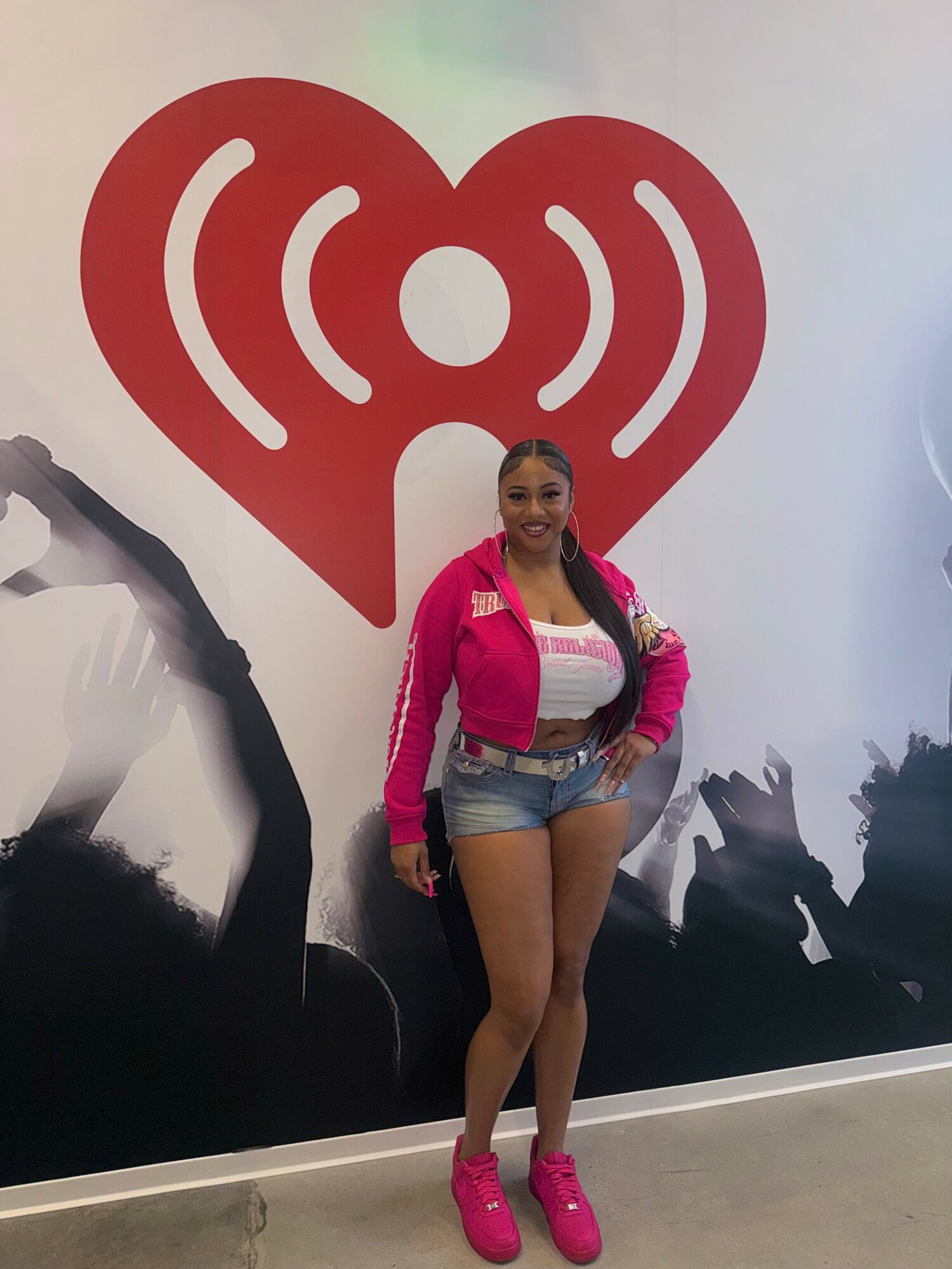 Woman standing in front of a large red heart with a Wi-Fi symbol, smiling, wearing pink shoes, denim shorts, and a pink jacket.