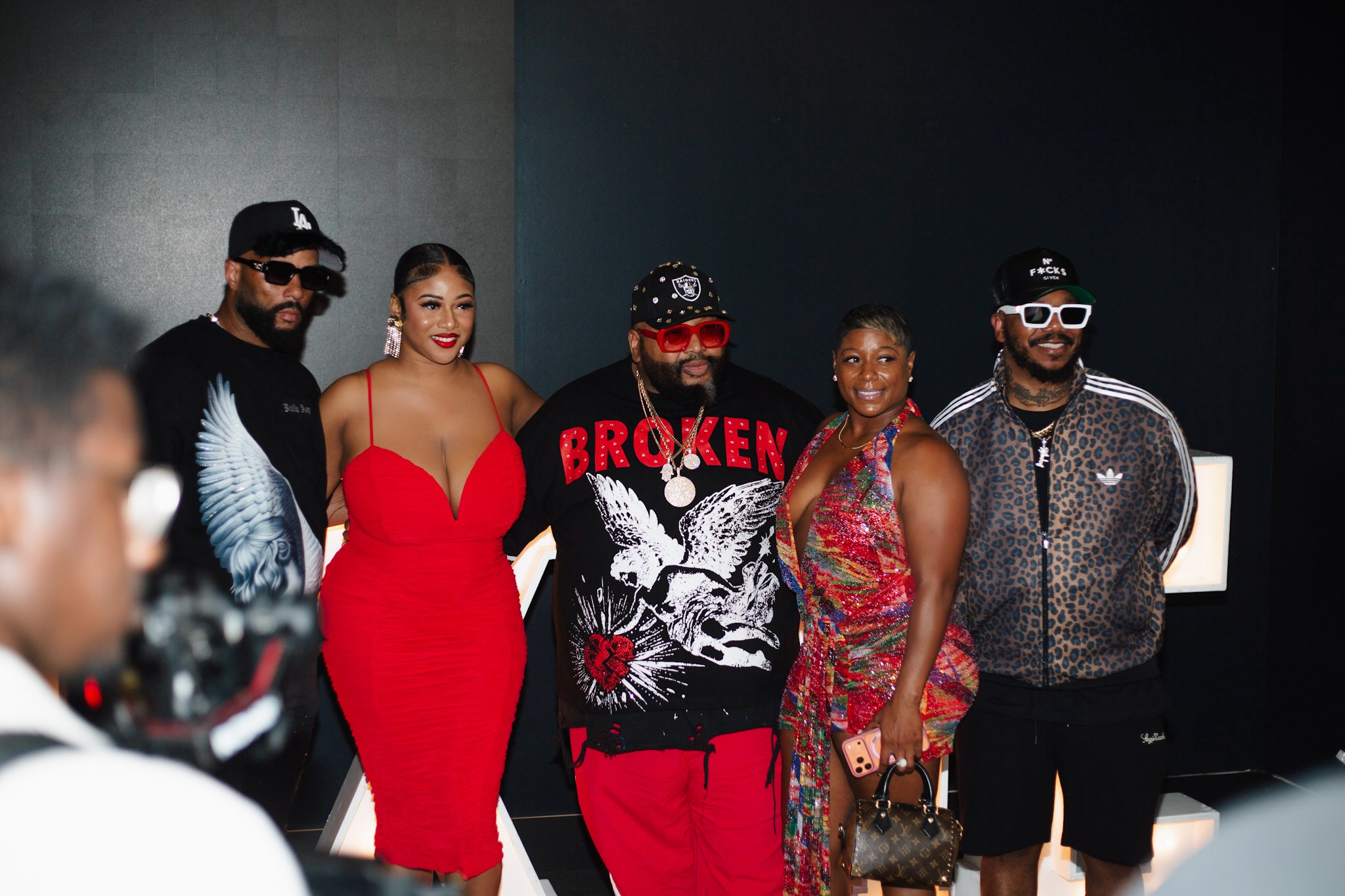 Group of five people standing together indoors, dressed stylishly, with a dark background.