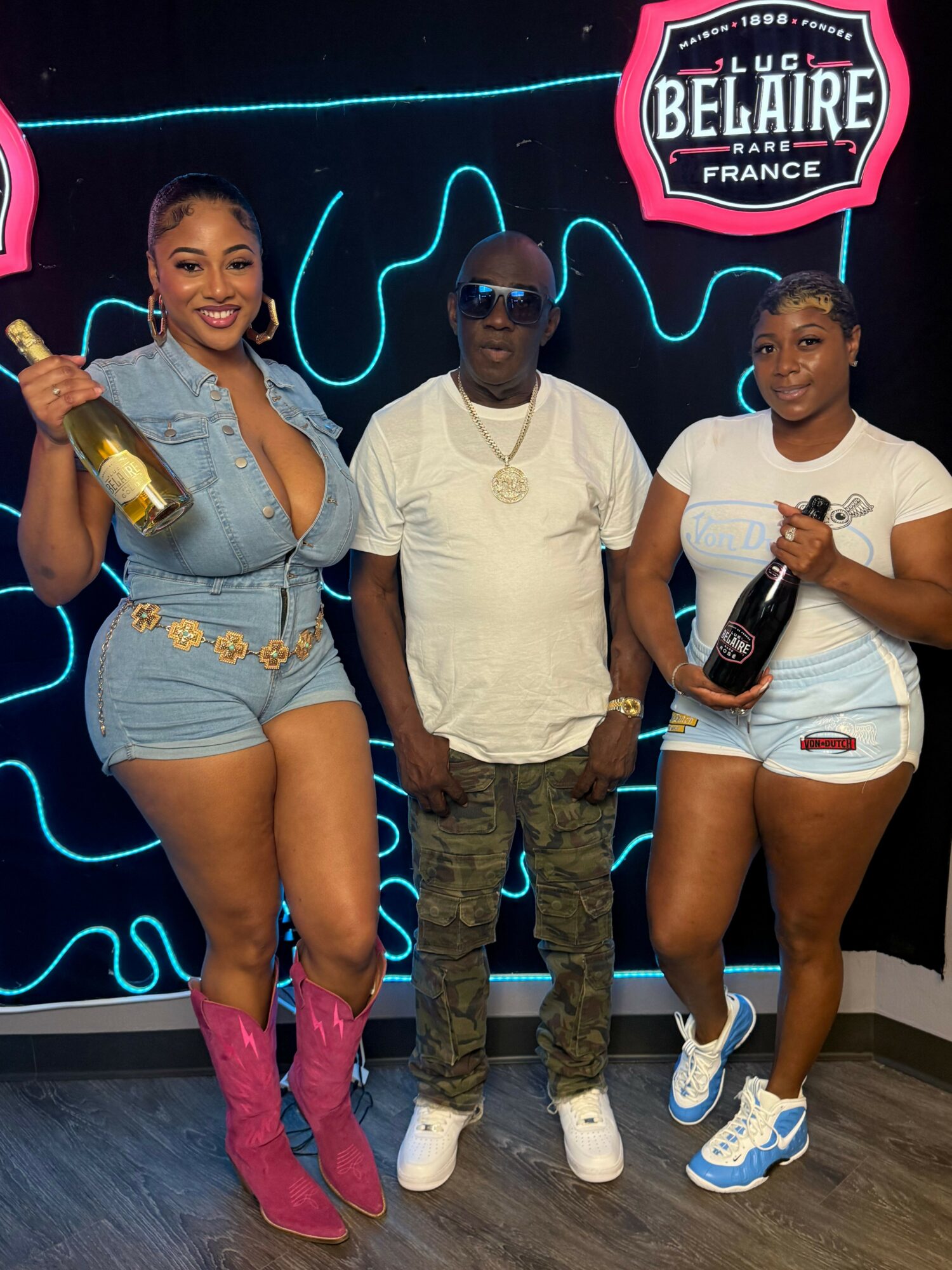 Three people posing in front of a neon sign, two women and one man, holding drinks and smiling.