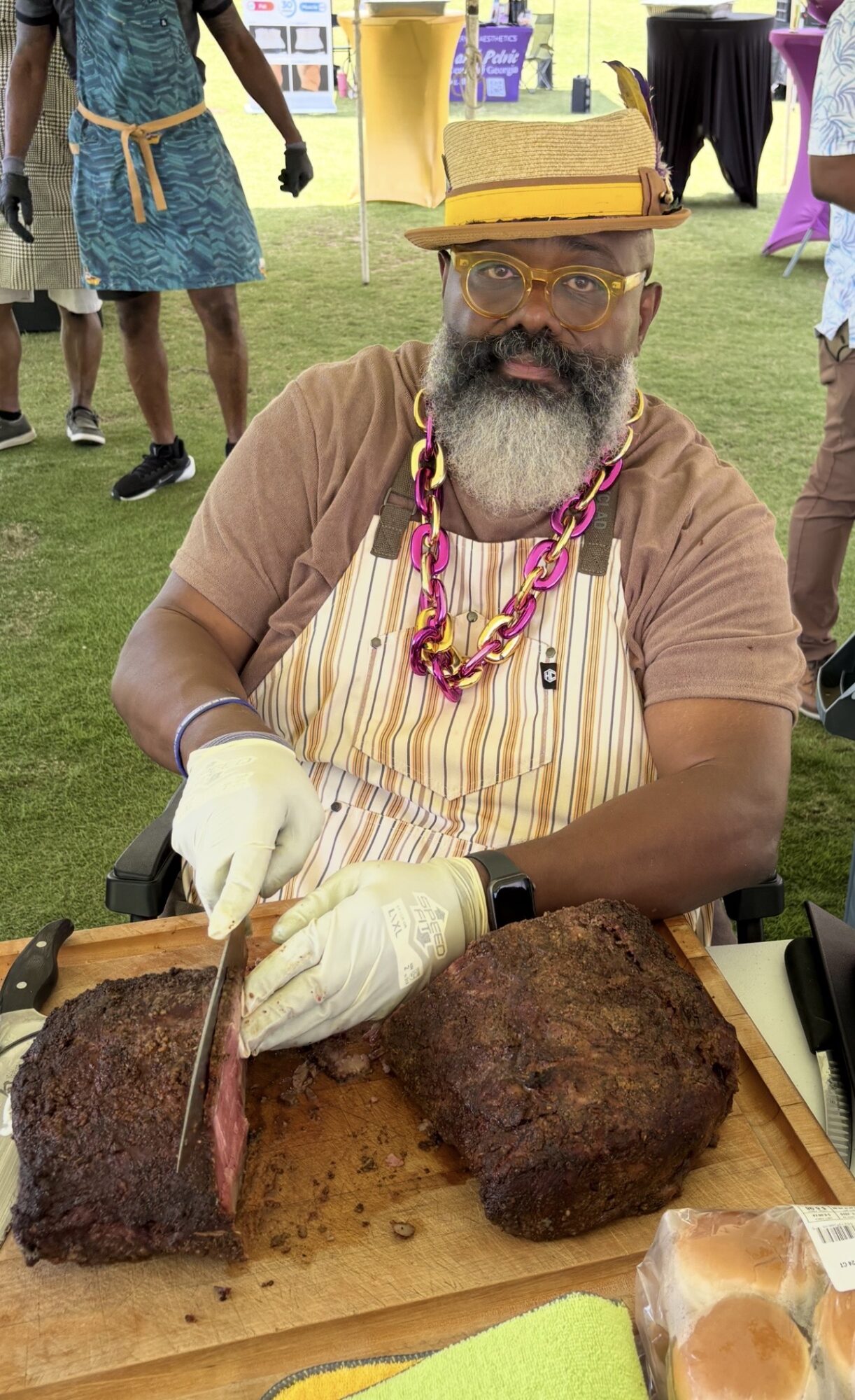 Man with glasses, beard, and colorful necklace slices meat at outdoor event, wearing apron and gloves.