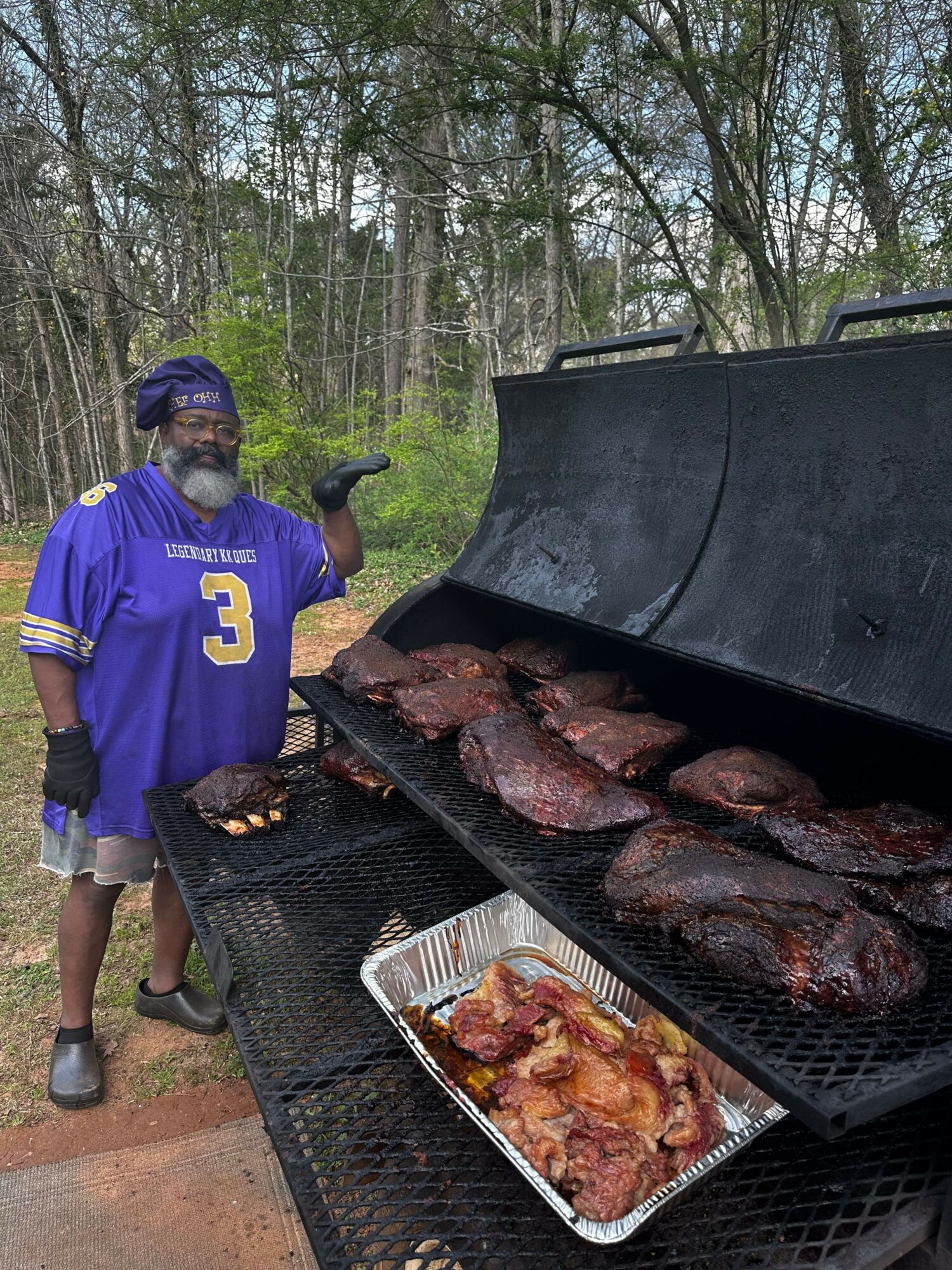 Person in a blue sports jersey and cap grilling meat outdoors with trees in the background.