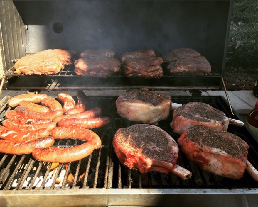 Grill with various meats and shrimp cooking, smoke rising, outdoors setting, food on grill racks.
