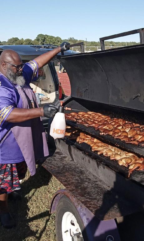 Person wearing a purple apron and gloves is grilling chicken on a barbecue with a lid open outdoors.