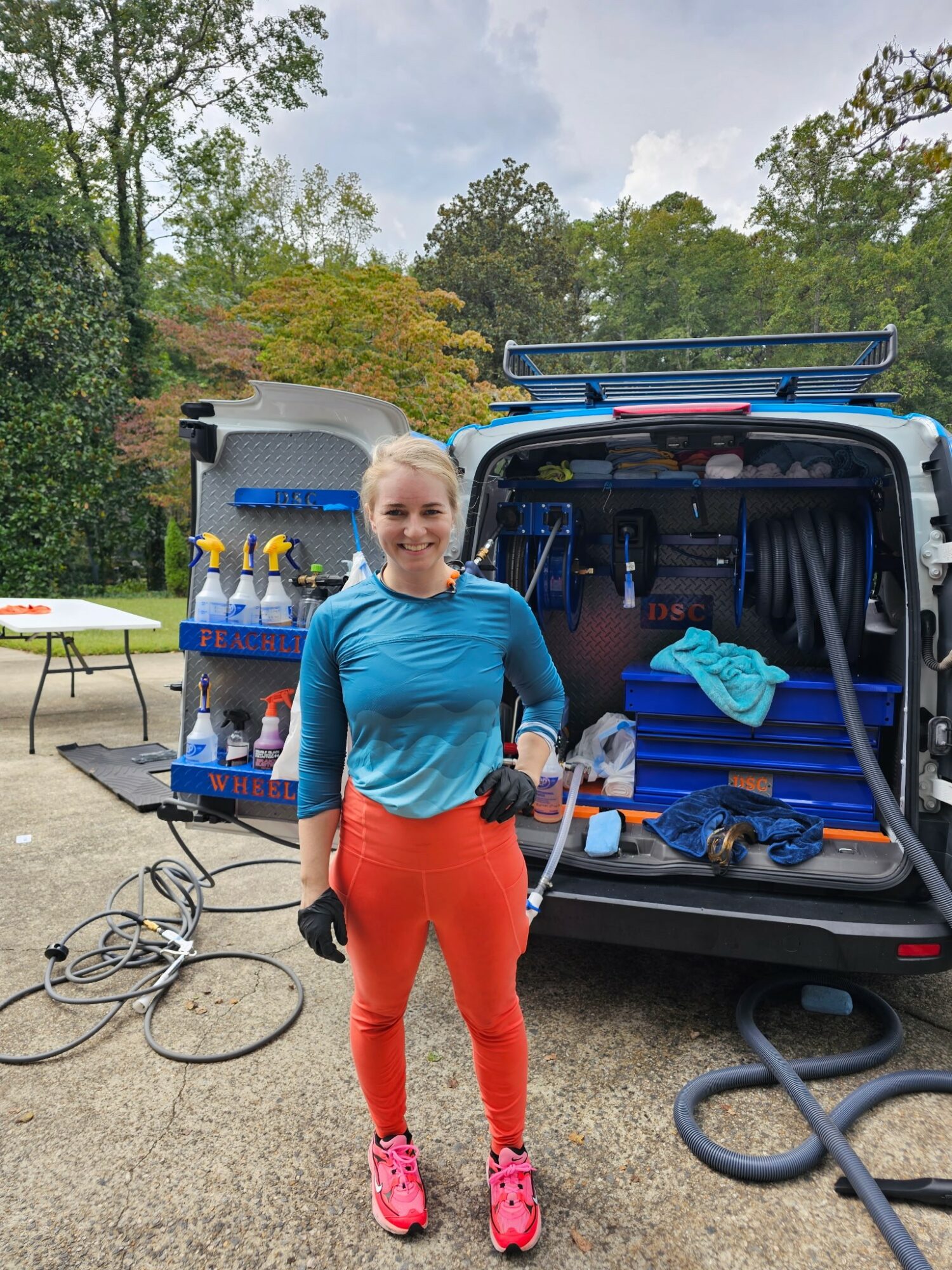 A woman in athletic clothing stands outdoors near a van with equipment, smiling, with trees and a cloudy sky in background.