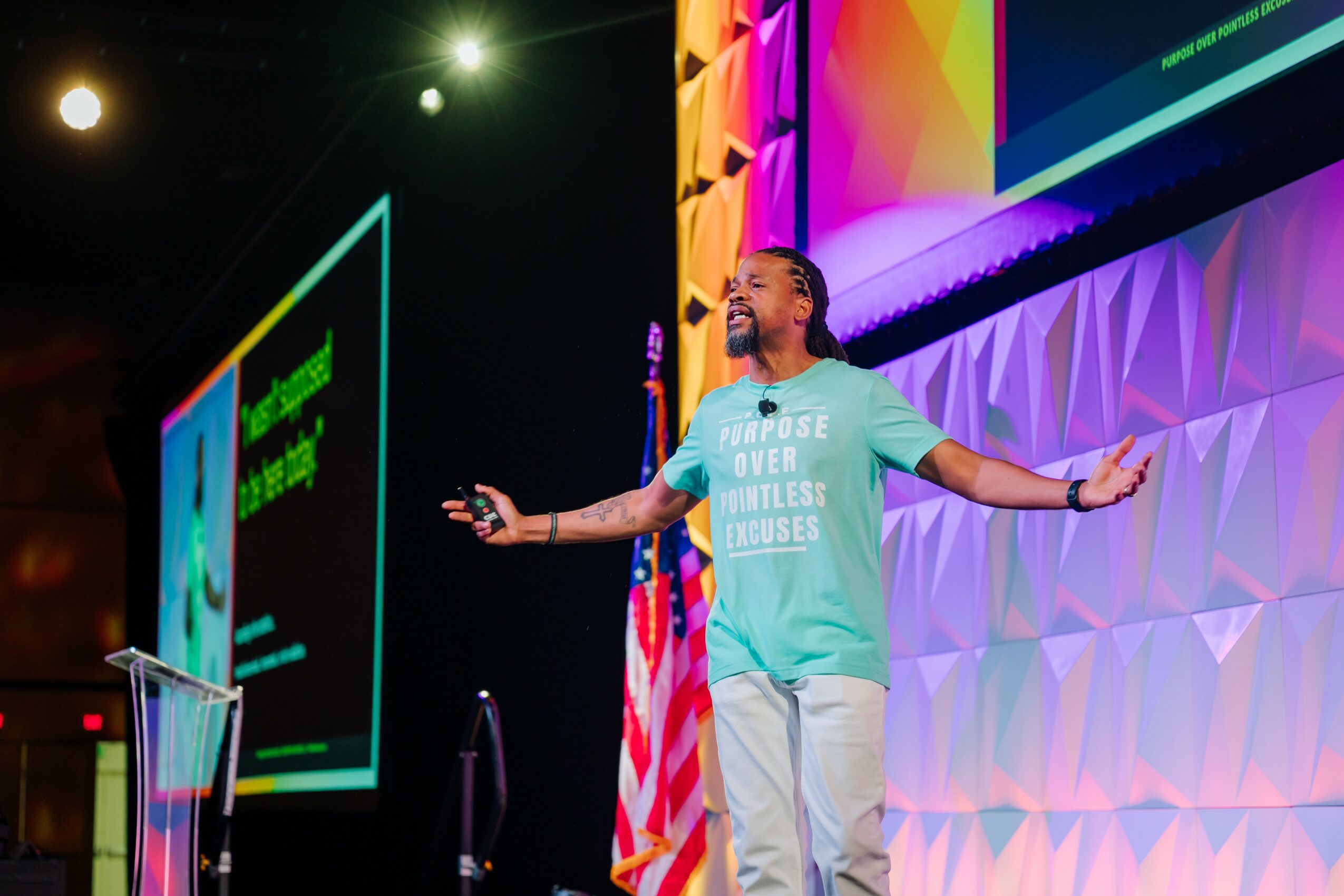 Person speaking on stage with colorful background and large screen, arms outstretched, wearing a light blue T-shirt and white pants.