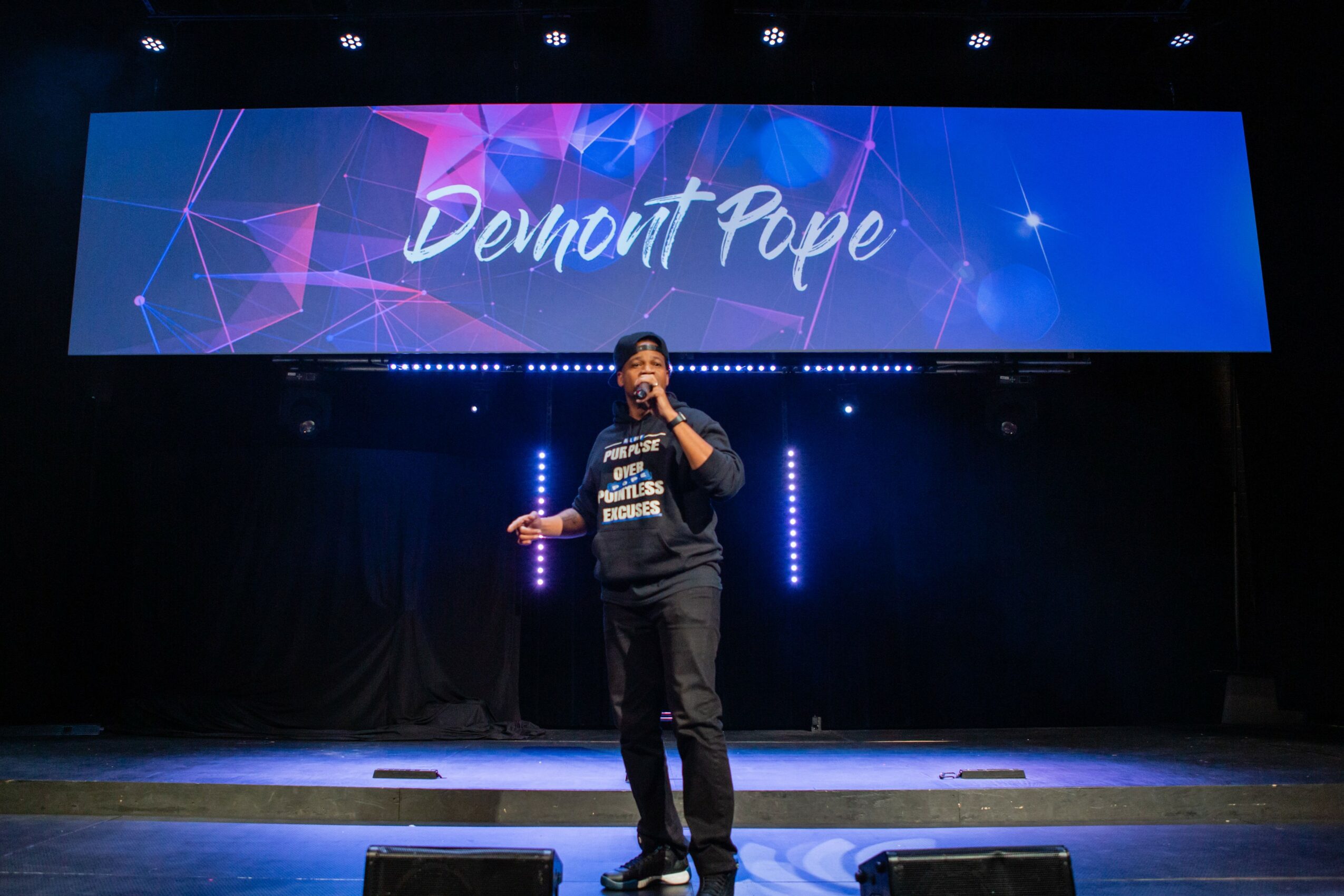 Person standing on stage holding a microphone, with a large screen behind displaying the name 'Demont Pope'.