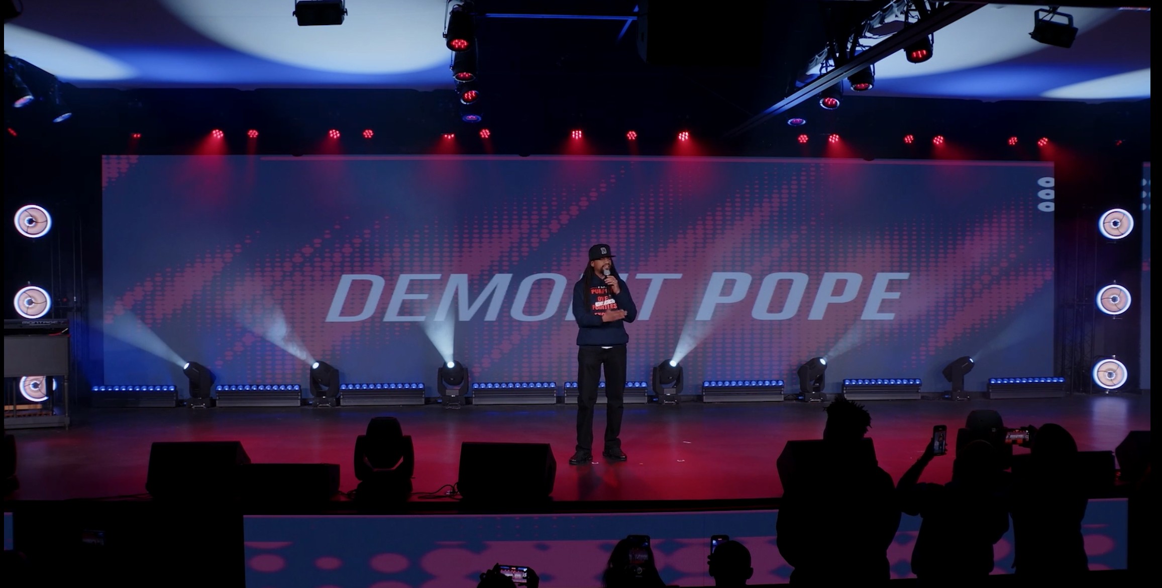 Person standing on stage with a large screen behind displaying 'DEMO AT POPE', audience silhouettes in foreground.