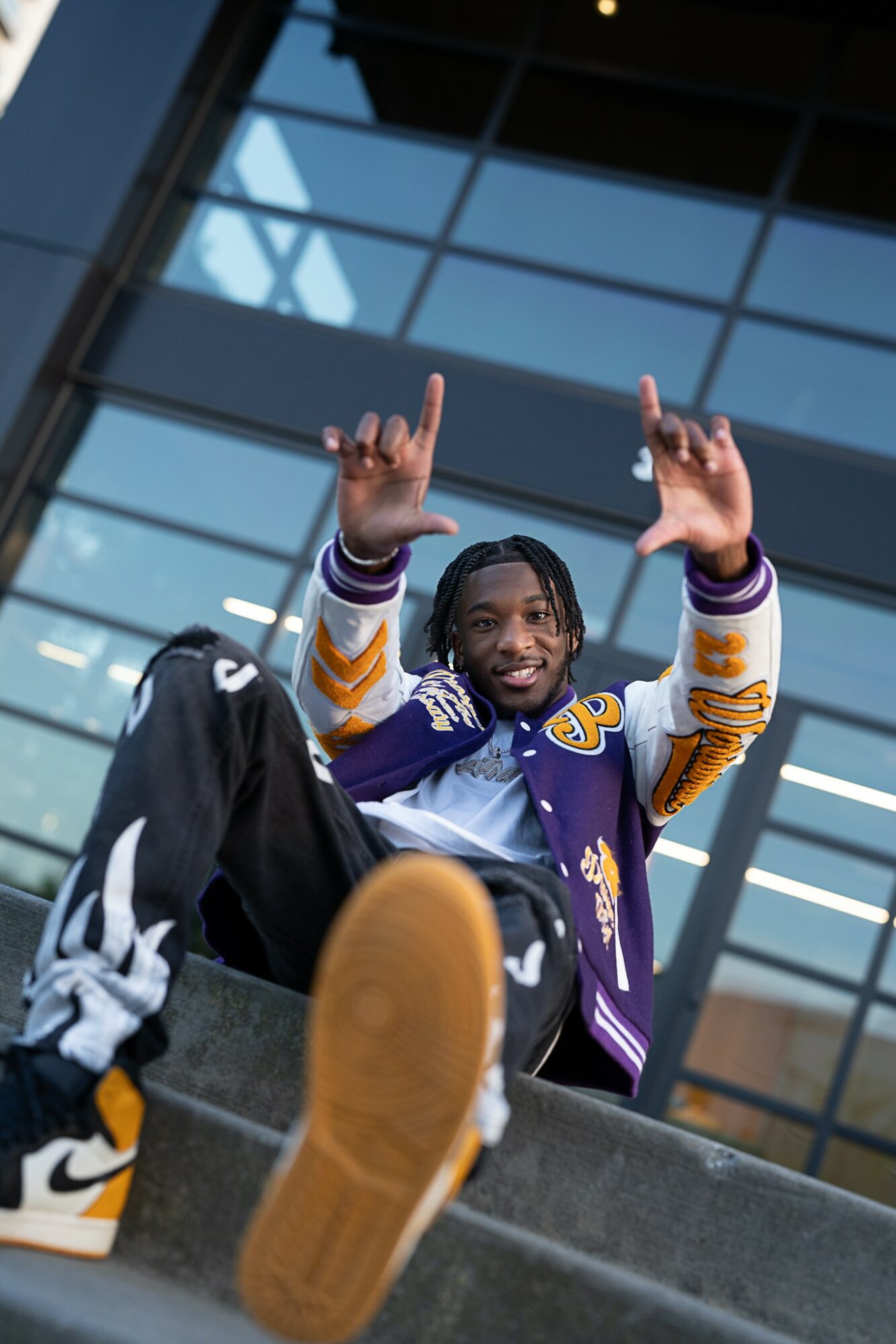 Young man sitting on stairs outside a modern glass building, smiling and raising both hands with peace signs.