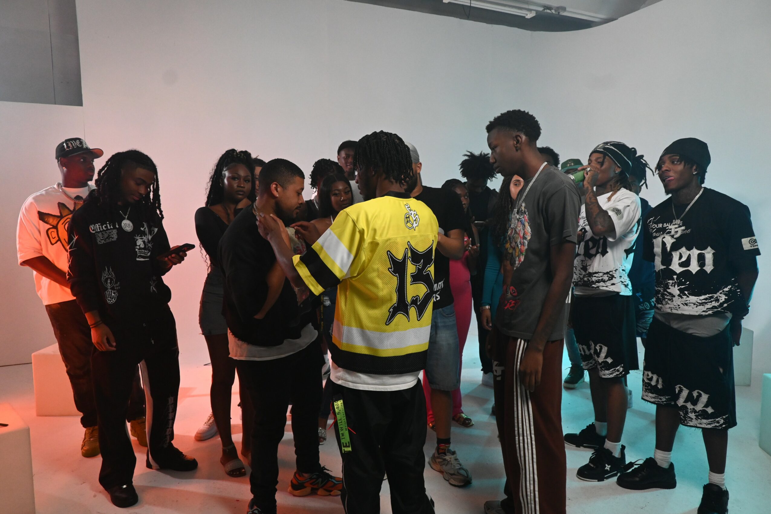 Group of people standing together indoors, some looking at a person in a yellow and black jersey, others talking or observing.