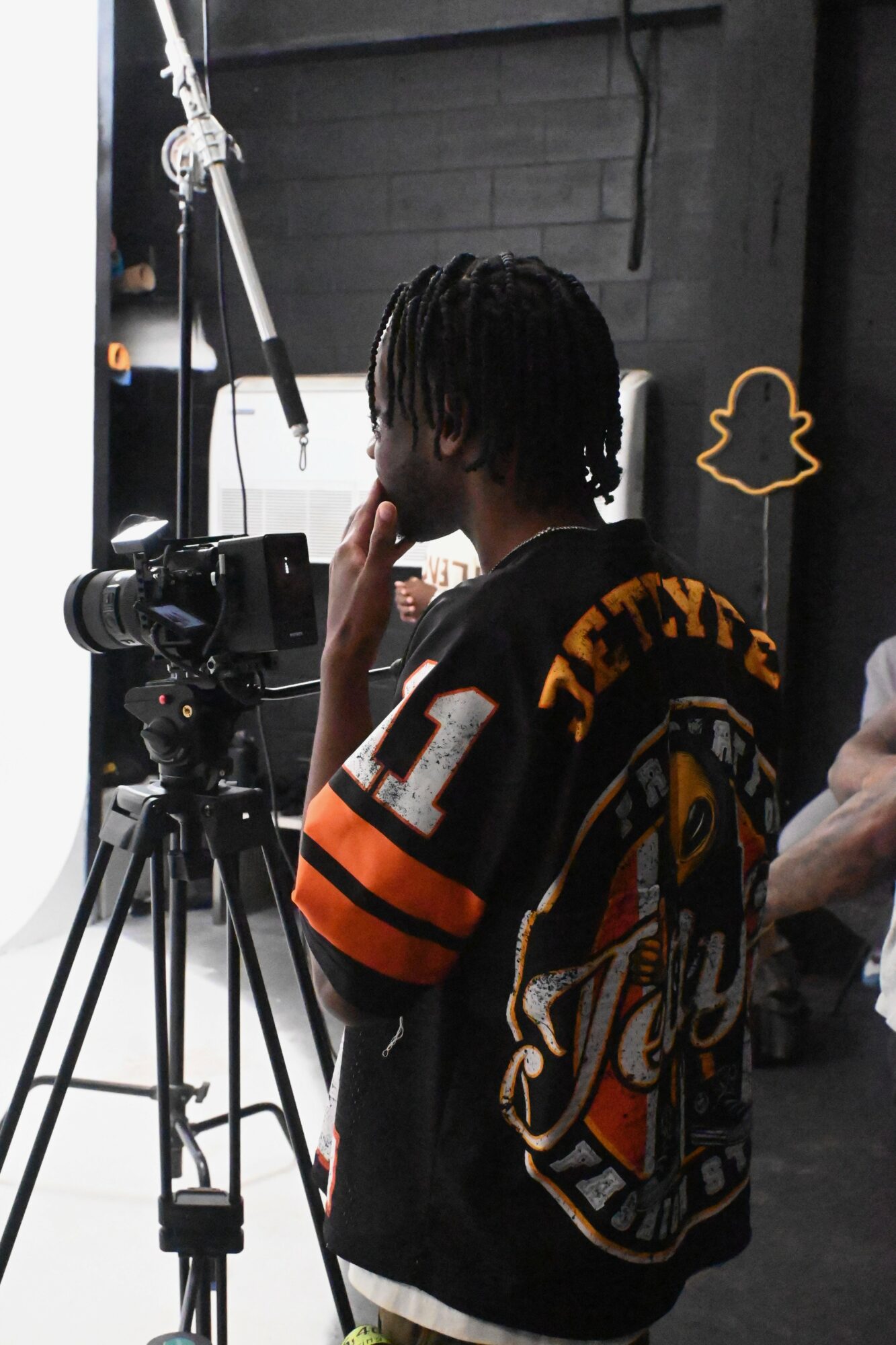 Person with dreadlocks wearing a black and orange sports jersey, standing next to a camera on a tripod in a studio.