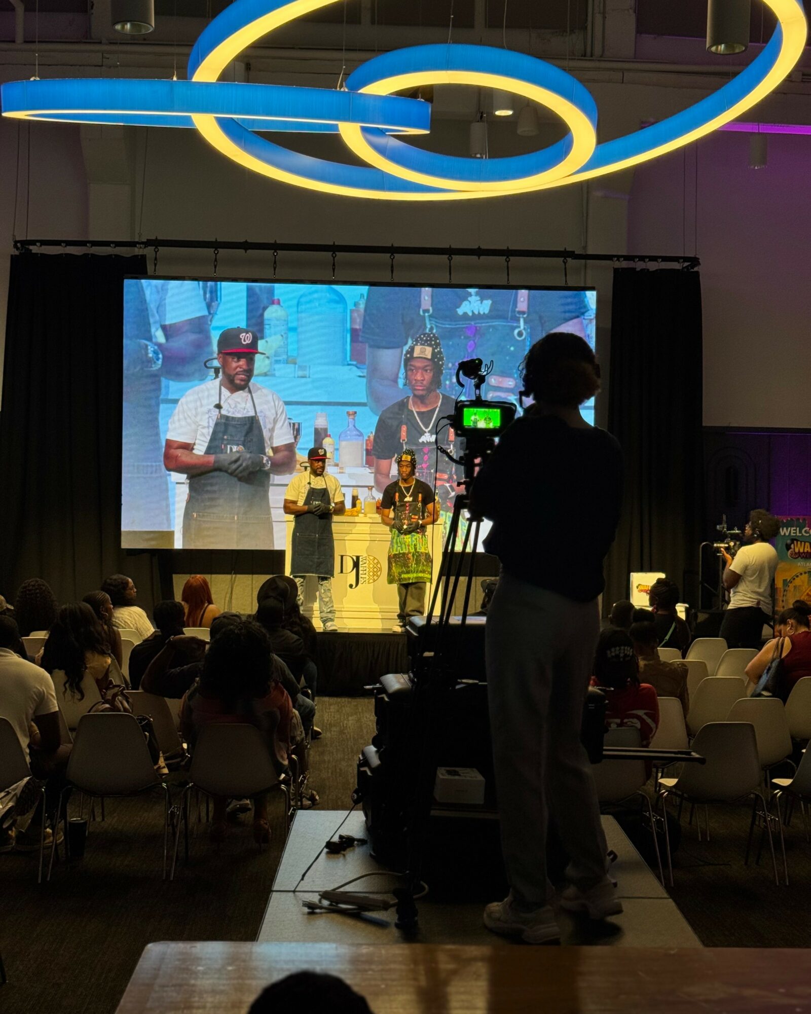 People on stage with a large screen behind them, audience seated in front, person filming in foreground, modern ceiling lights above.