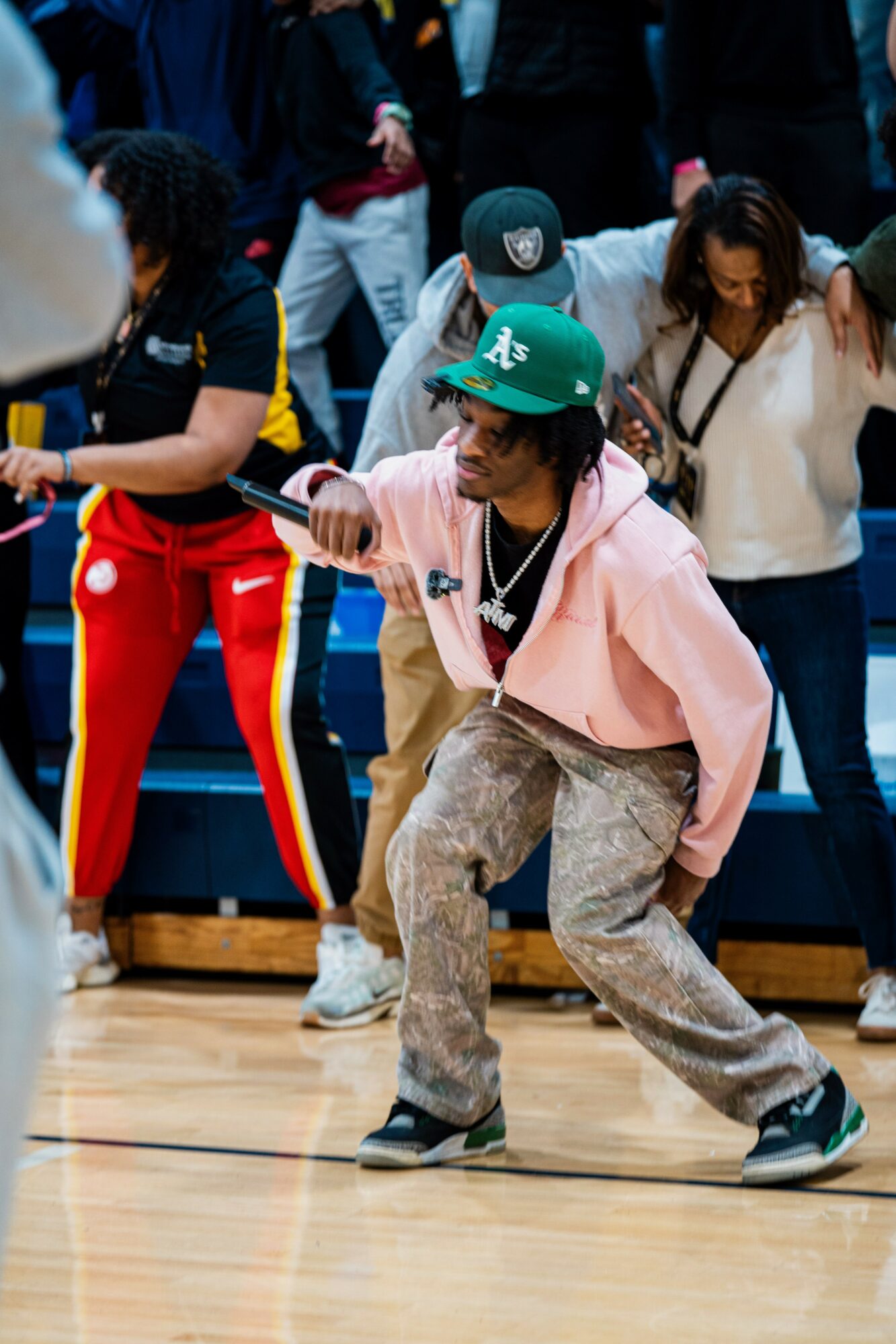 Person wearing a green cap, pink hoodie, and patterned pants dancing on a gym floor, surrounded by people.