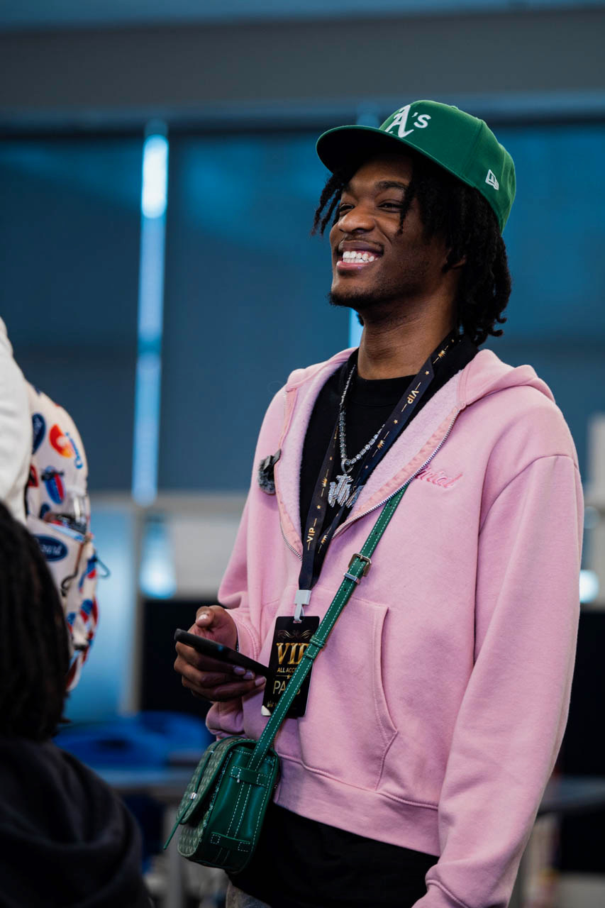 Young man with dreadlocks wearing a green cap, pink hoodie, and holding a phone, smiling.