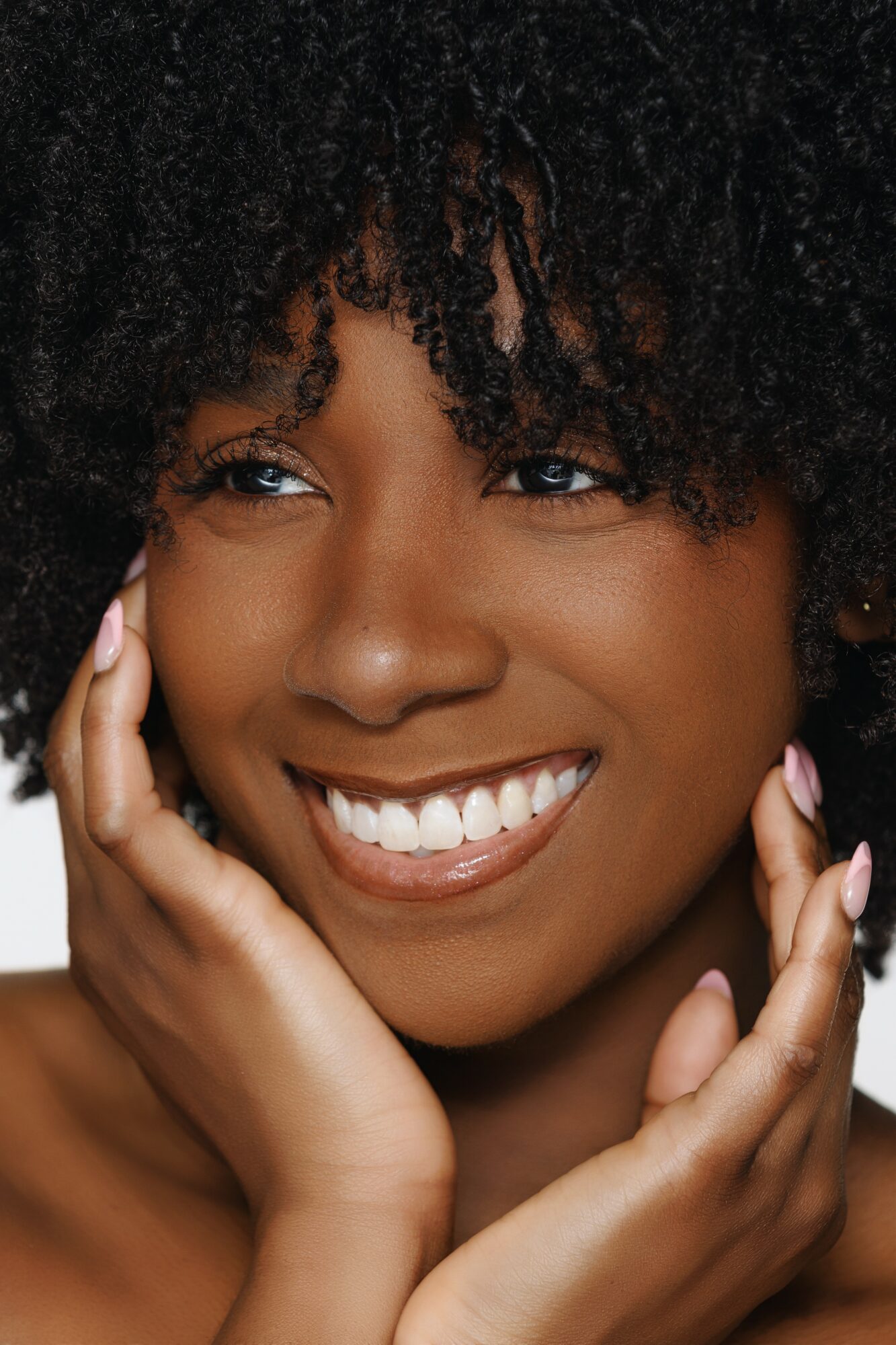 Close-up of a smiling woman with curly hair, touching her face with both hands.