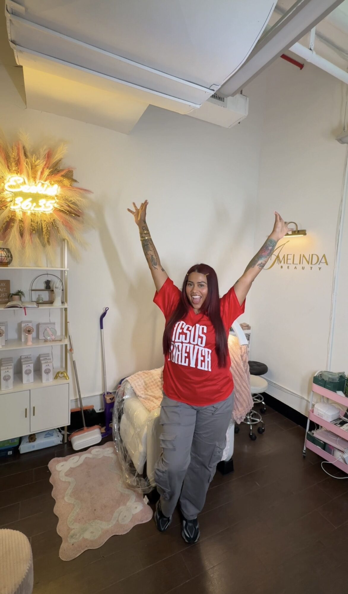 Person with arms raised in a room with a white wall, shelves, and a pink cart, wearing a red shirt and gray pants.
