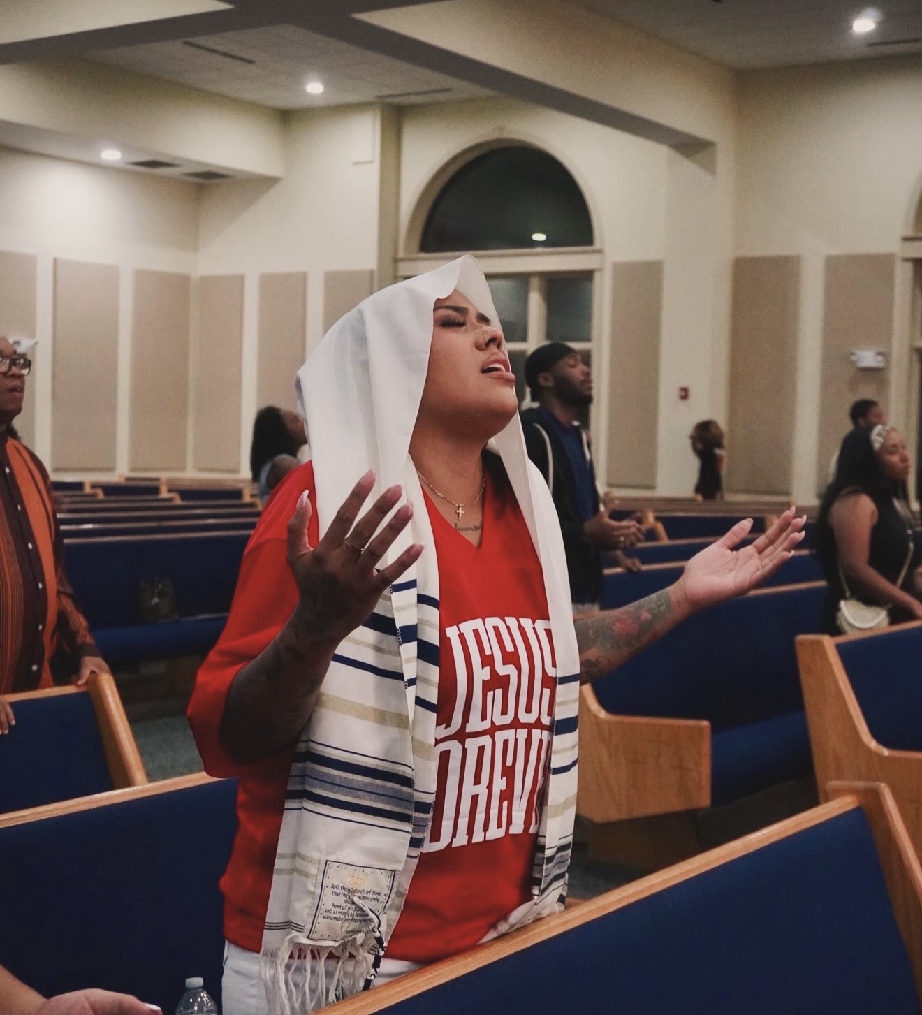 Woman with a white head covering and red shirt praying with hands raised in a church, others in background.