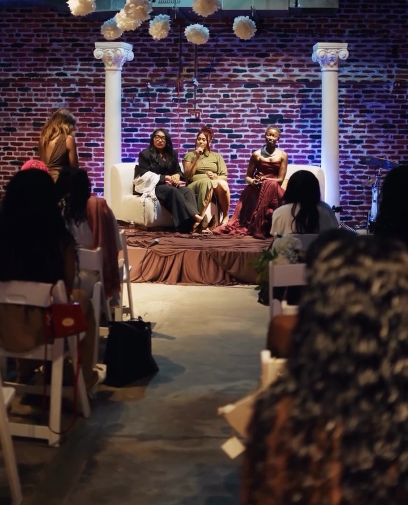 Four women sit on a stage with a brick wall background, columns, and hanging decorations, speaking to an audience.