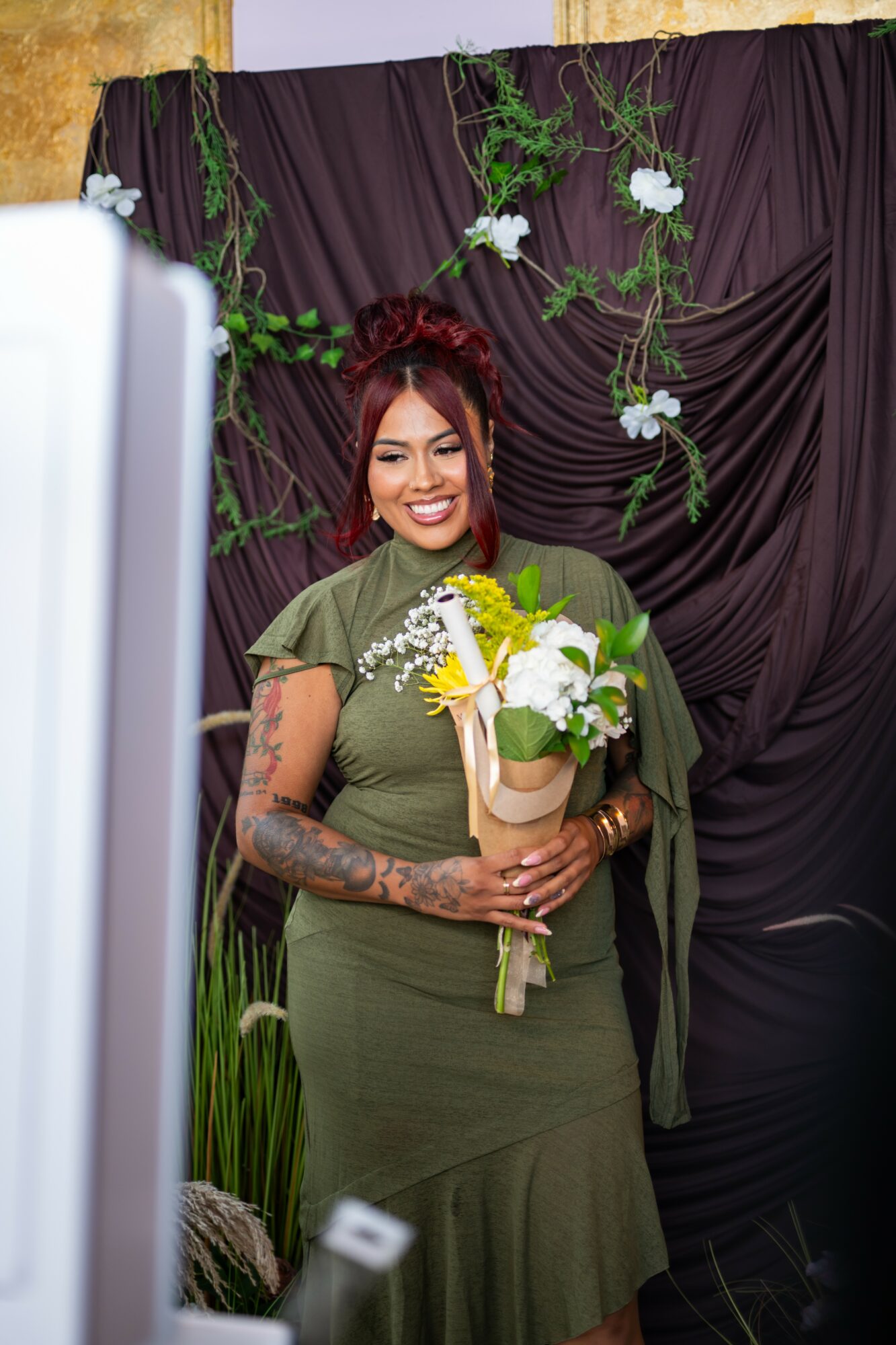Woman with red hair holding a bouquet of yellow and white flowers, standing in front of a dark curtain with green plants and white flowers.