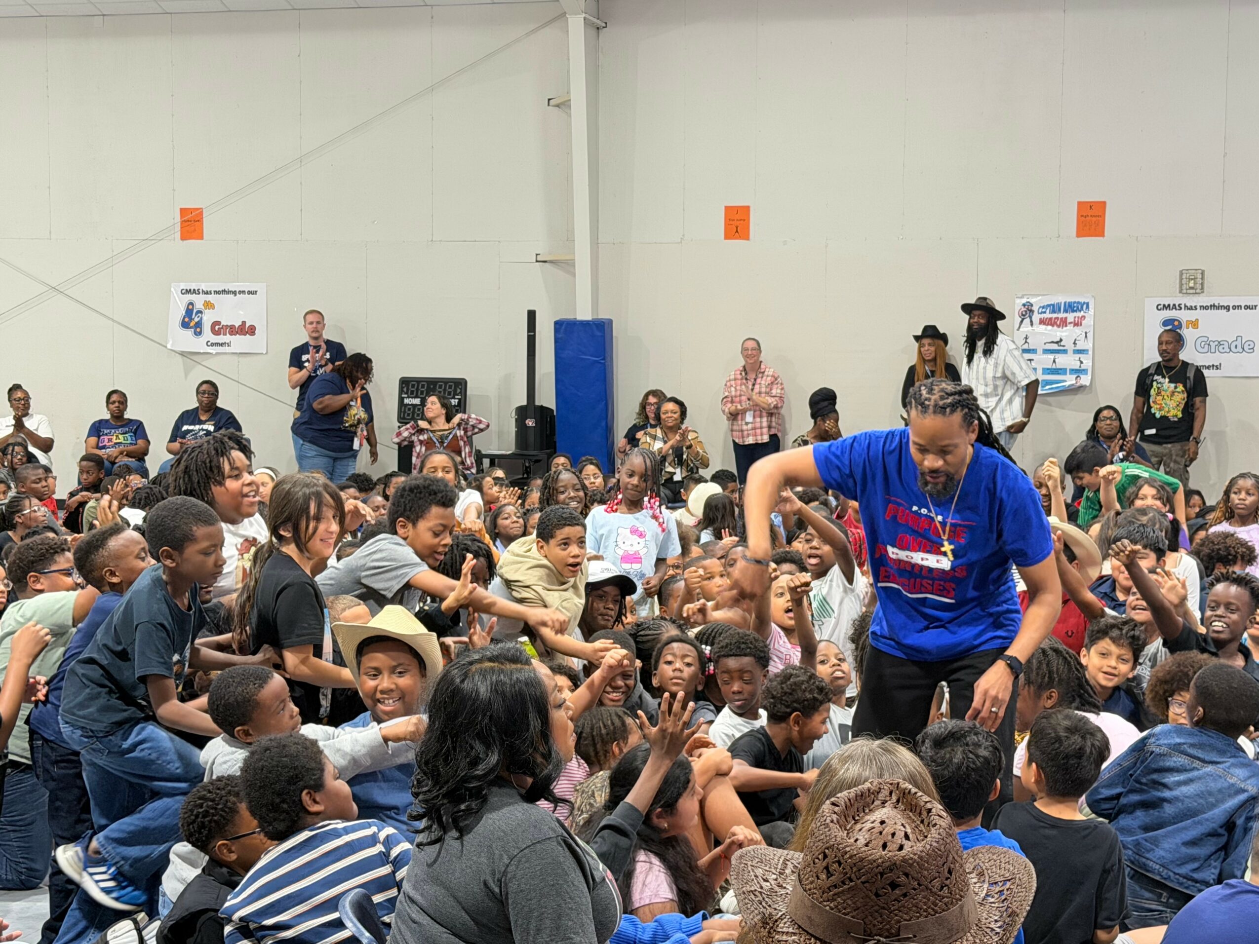 A woman in a blue shirt interacts with children sitting on the floor in a large indoor space, with many people watching.