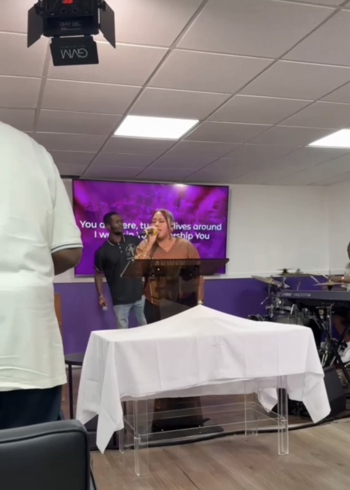 People standing in front of a purple screen with text, in a room with a white table and ceiling lights.