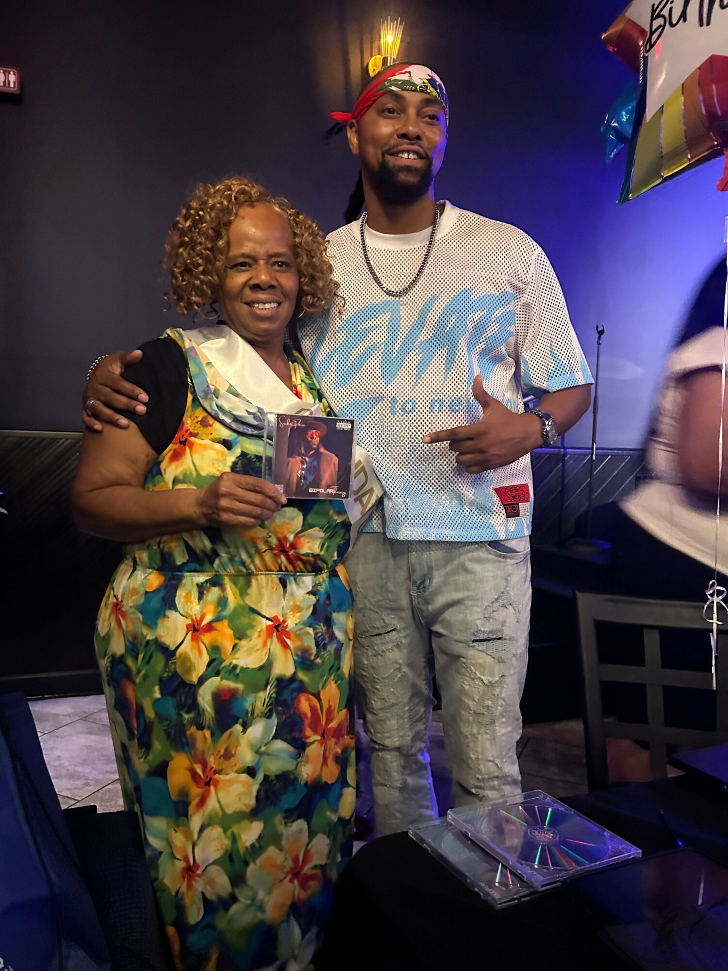 Two people posing indoors, one wearing a floral dress and the other in a light-colored outfit, with a dark background.