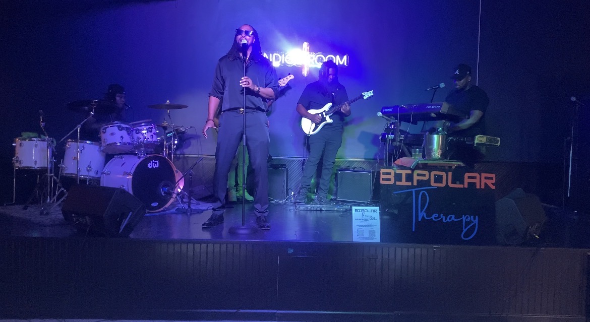 Band performs on stage with four musicians, one singing, others playing instruments, in a dimly lit setting with a sign reading 'BIPOLAR' behind them.