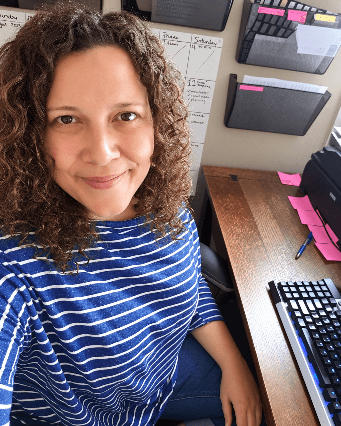 Woman with curly hair smiling in a striped shirt at a desk with office supplies and a keyboard.