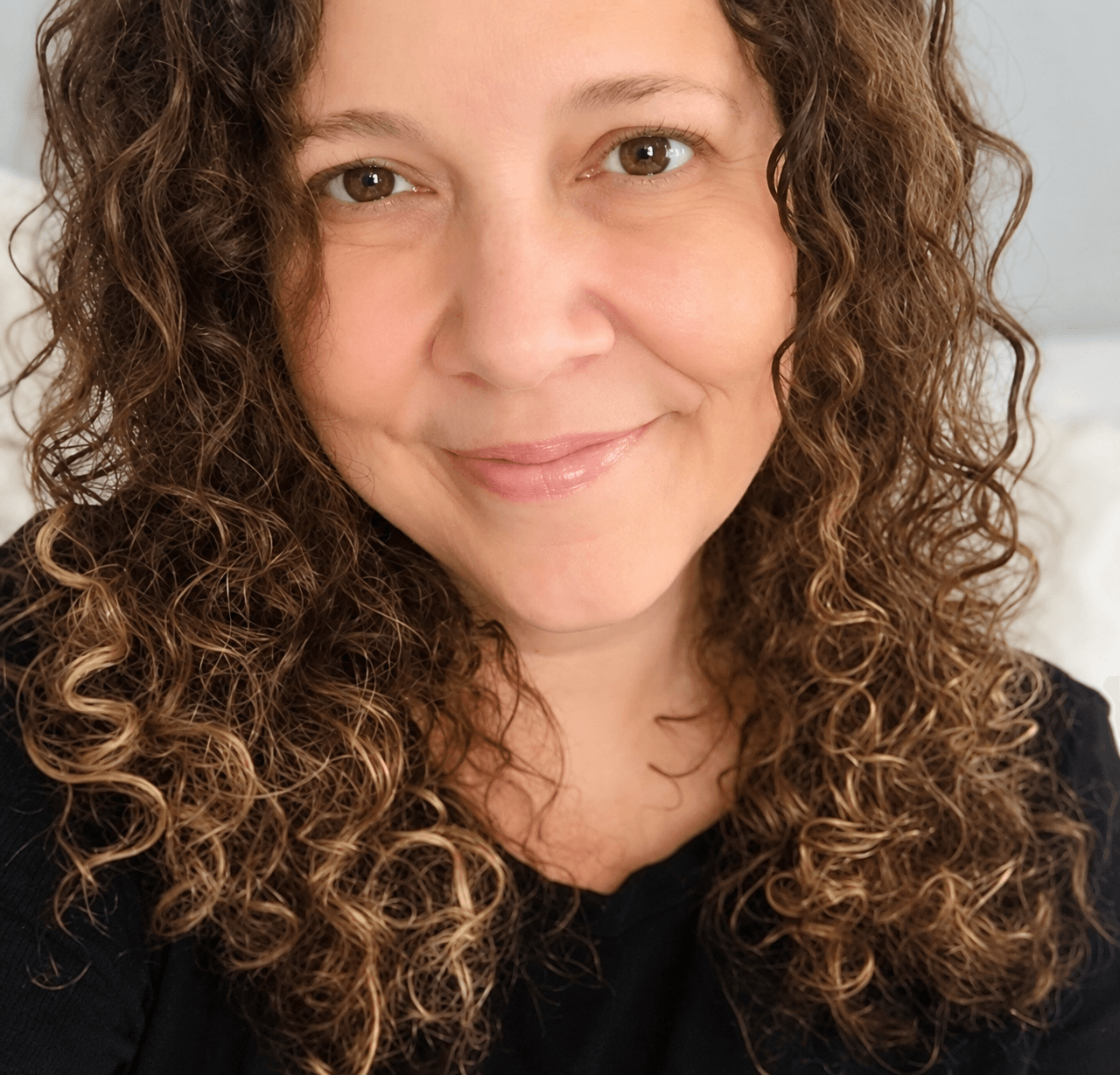 Close-up of a woman with curly hair smiling, wearing a black top, against a light background.