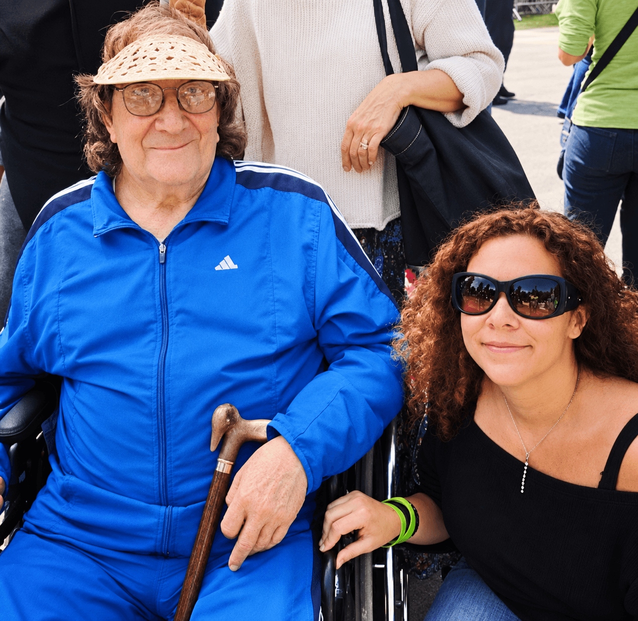 Two women outdoors, one elderly with glasses and a hat, the other younger with sunglasses, smiling together.