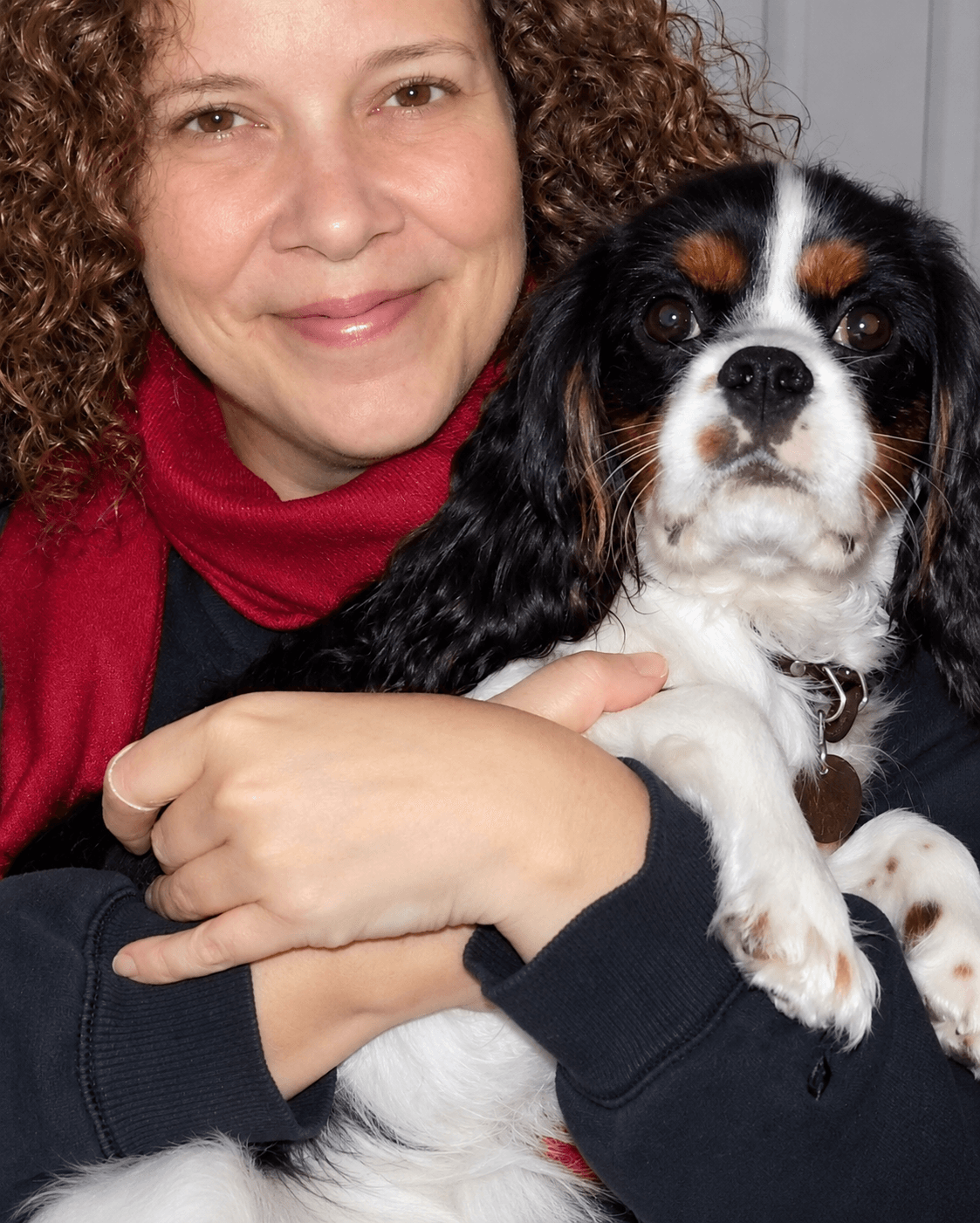 Person with curly hair smiling, holding a small black, white, and brown dog, indoors.