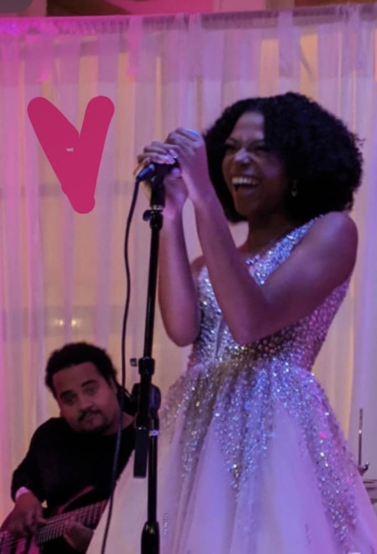 Smiling woman in a sparkly dress holding a microphone, with a man playing guitar nearby, in front of pink curtains.