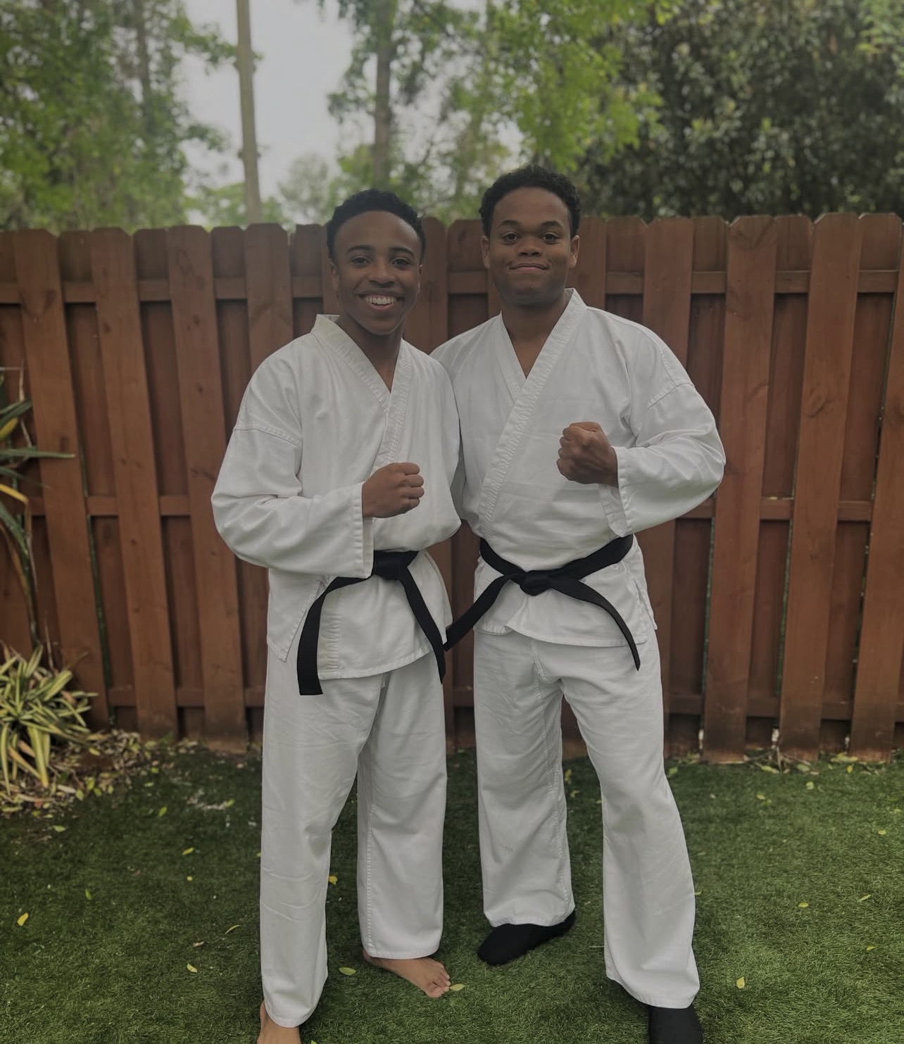 Two people in martial arts uniforms with black belts standing outdoors in front of a wooden fence, smiling and posing with fists raised.