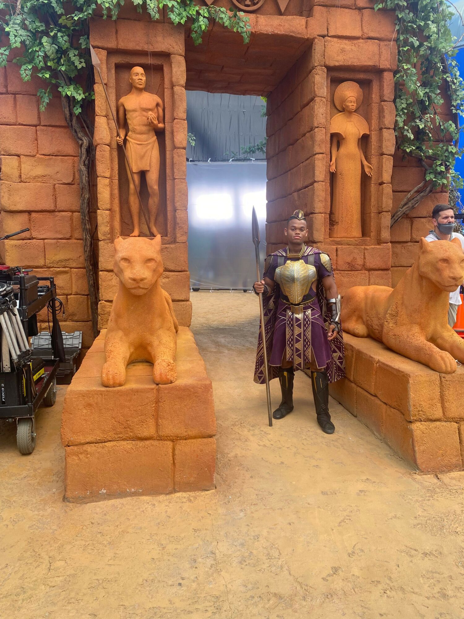 Two stone statues of seated figures in front of a brick archway, with a person dressed as a skeleton in costume nearby.