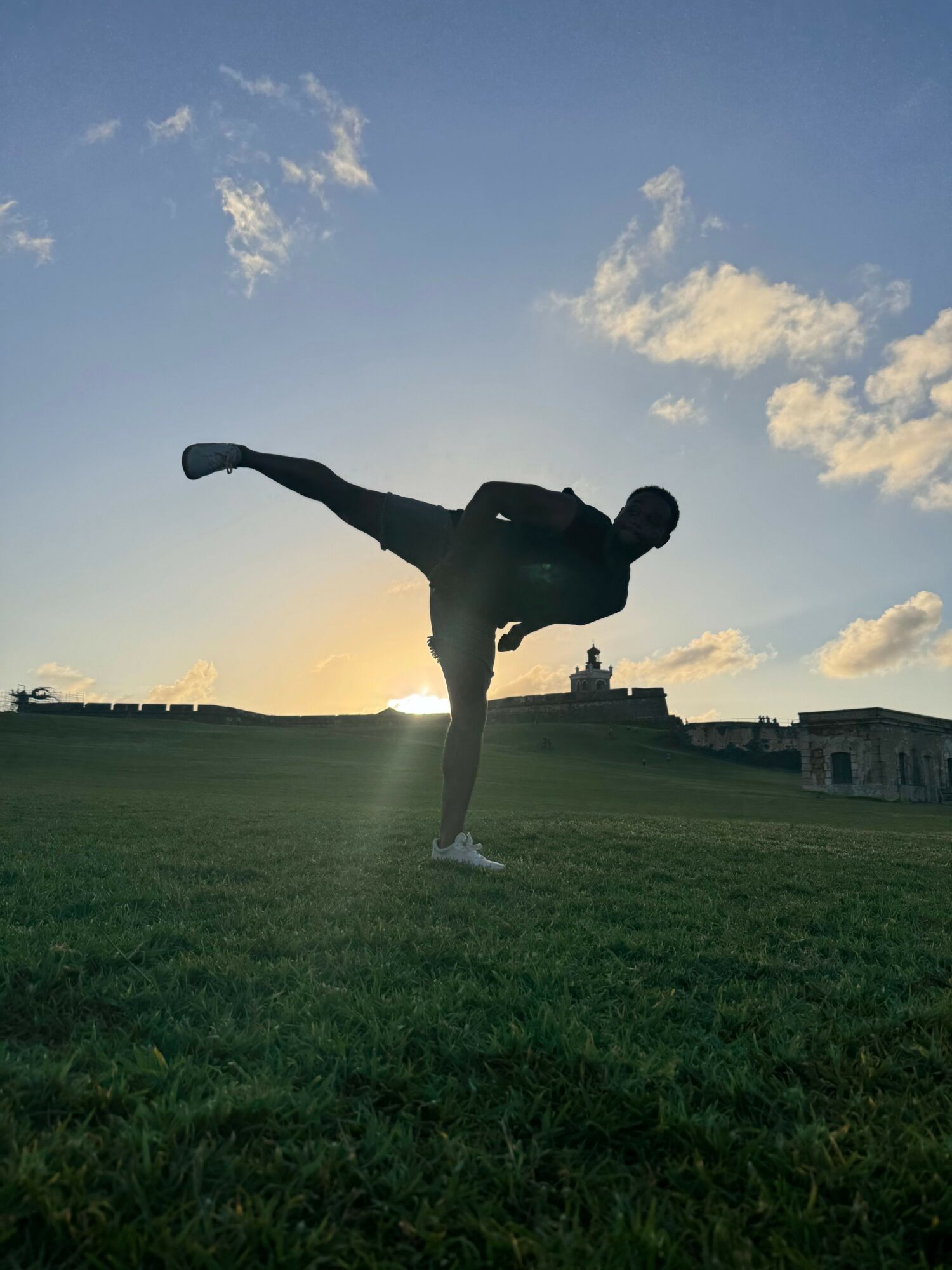Silhouette of a person jumping with arms and legs spread, sunset sky with clouds in the background, grassy field.