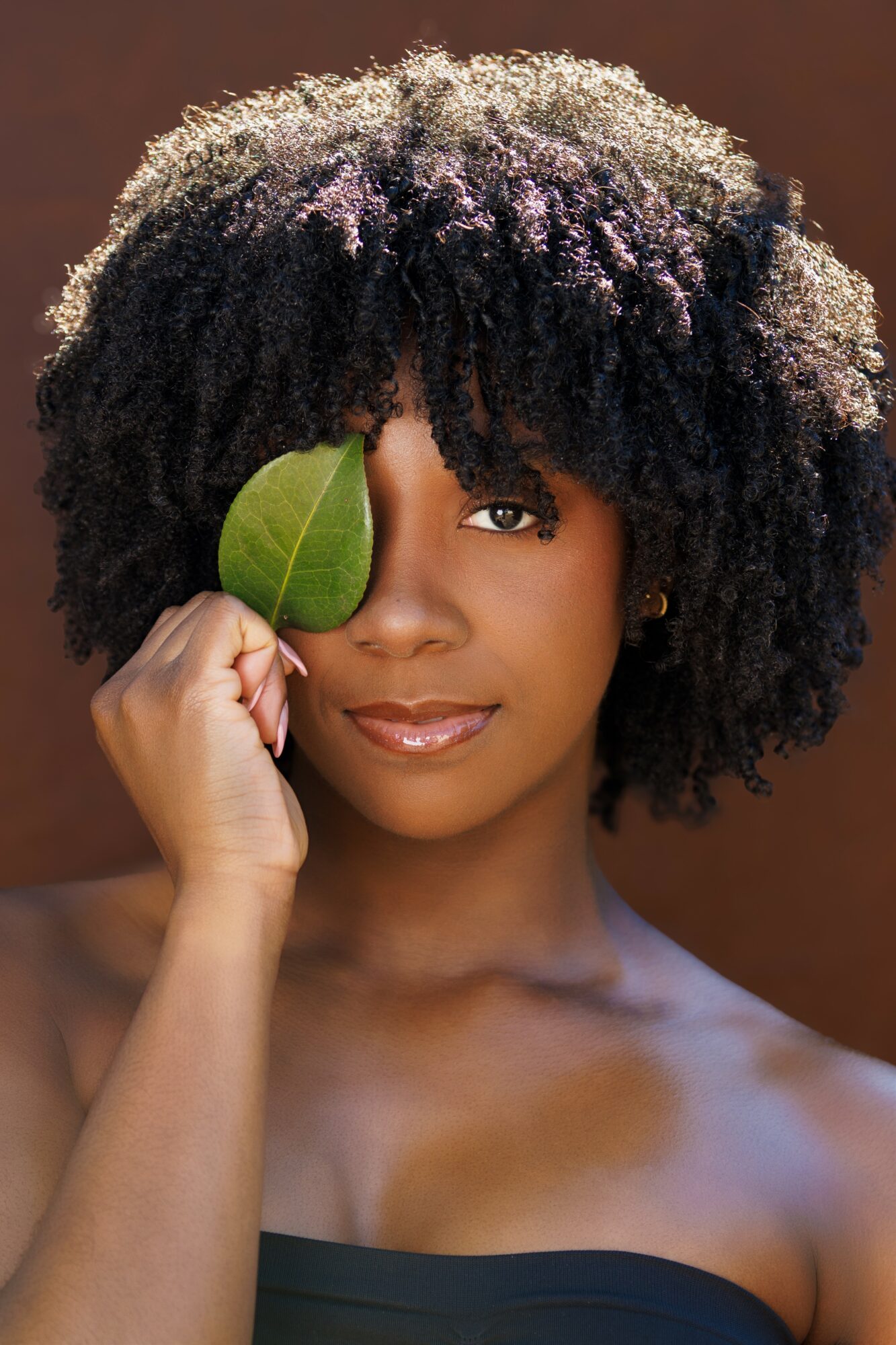 Woman with curly hair holds a leaf over her eye, facing forward, with a neutral background.