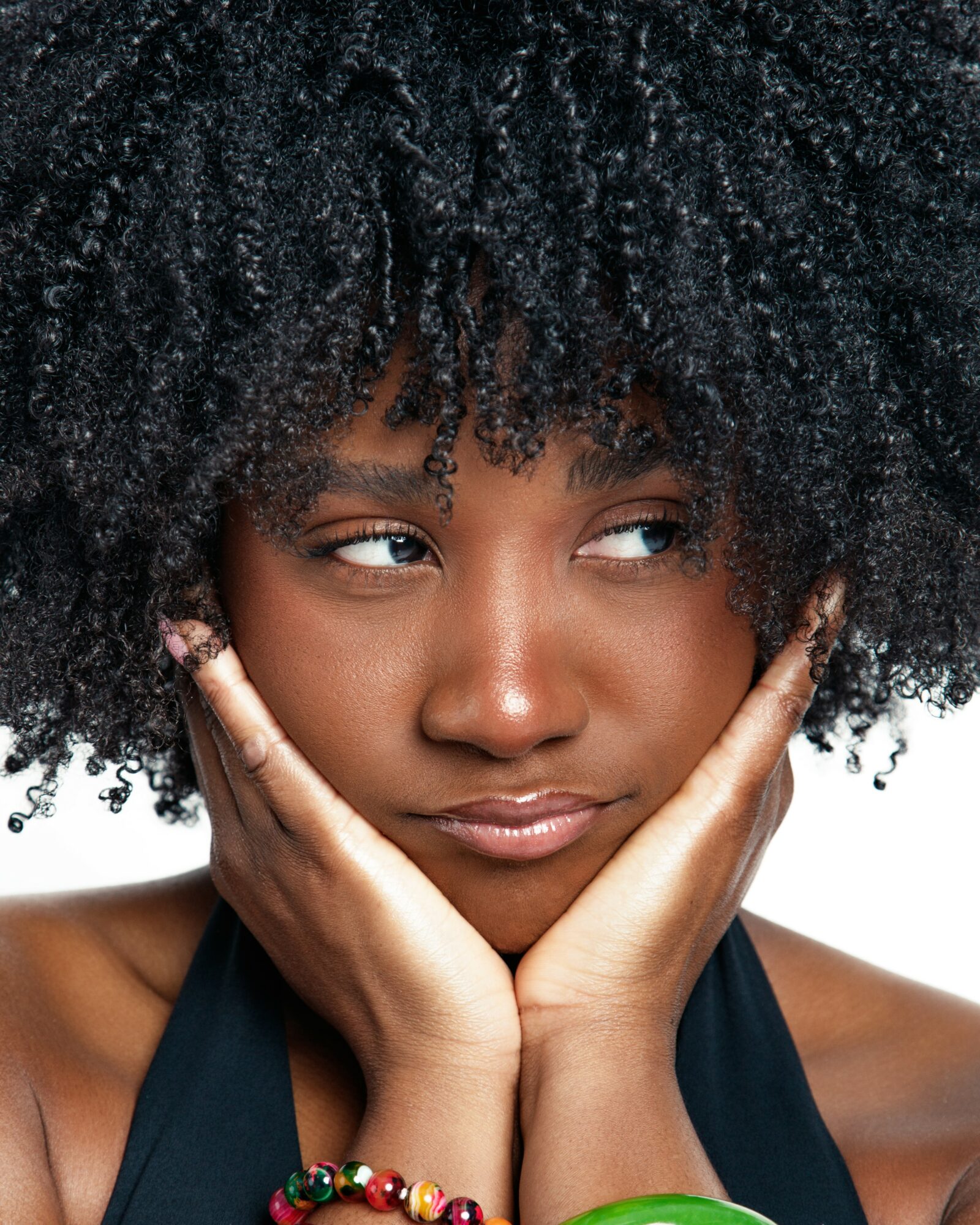 Young woman with natural curly hair resting her face in her hands, wearing a black top, against a white background.