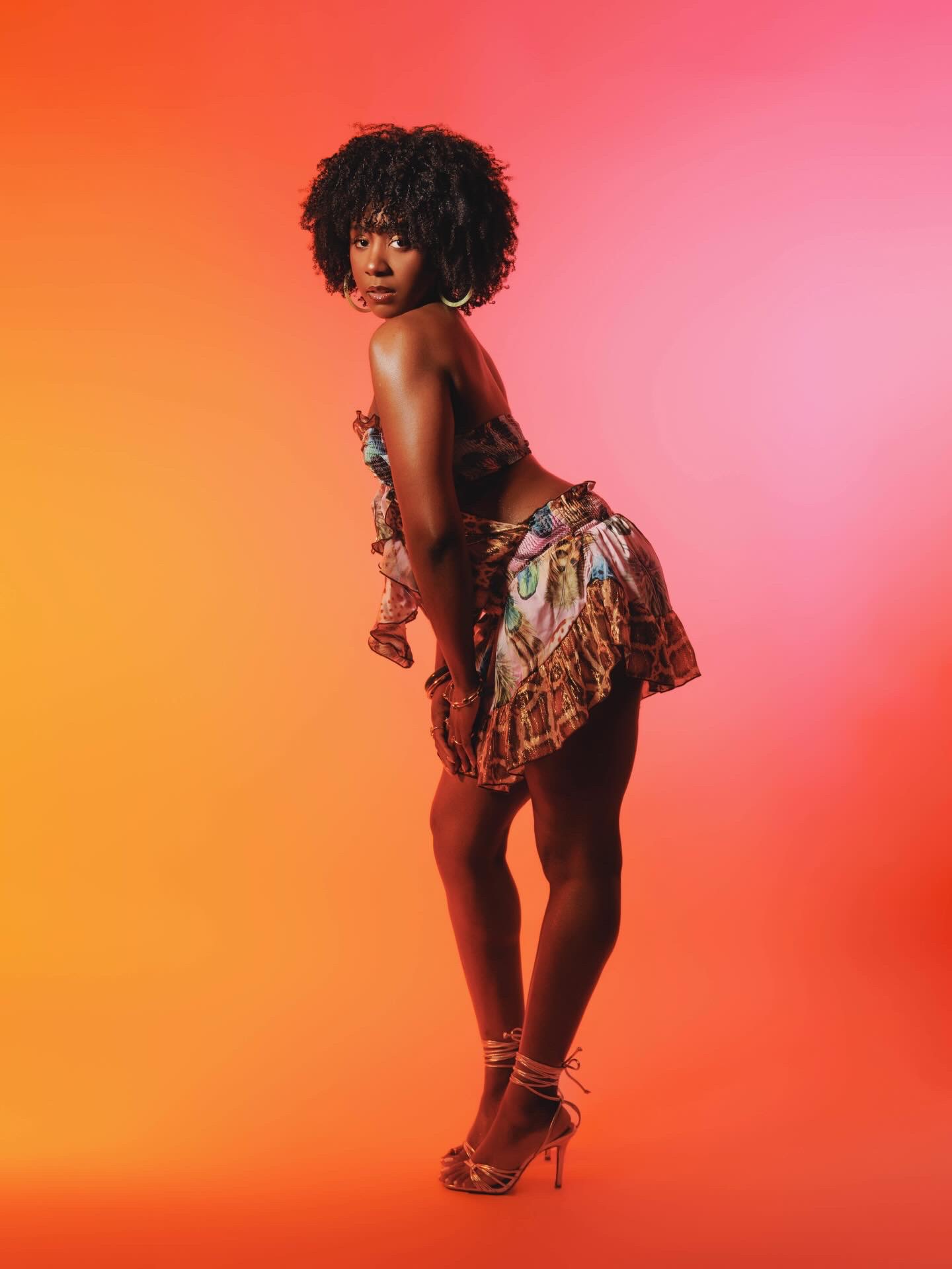 Young woman with curly hair in a colorful dress, standing against a pink and orange gradient background.