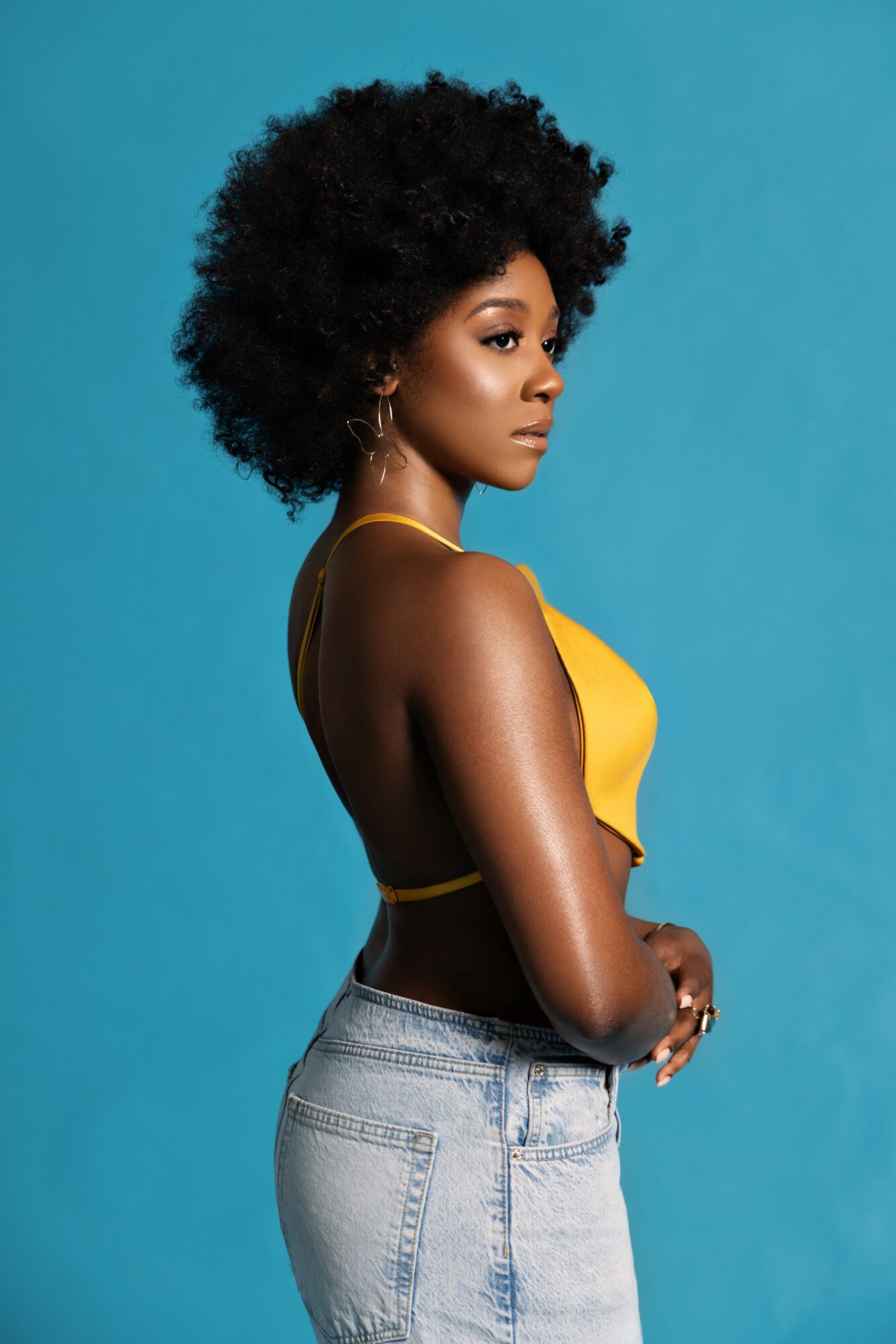 Side profile of a woman with natural curly hair wearing a yellow top and light jeans against a blue background.