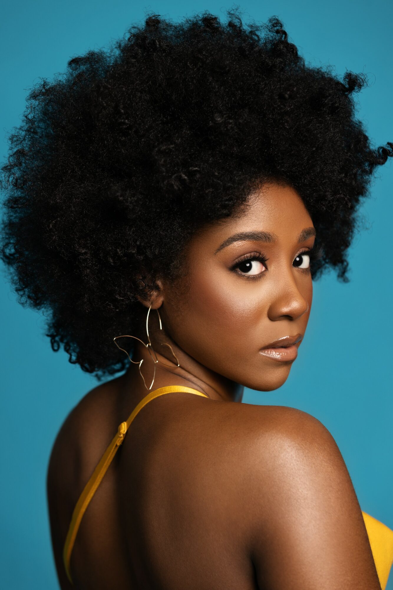 Portrait of a woman with natural curly hair, wearing hoop earrings and a yellow strap top, against a blue background.