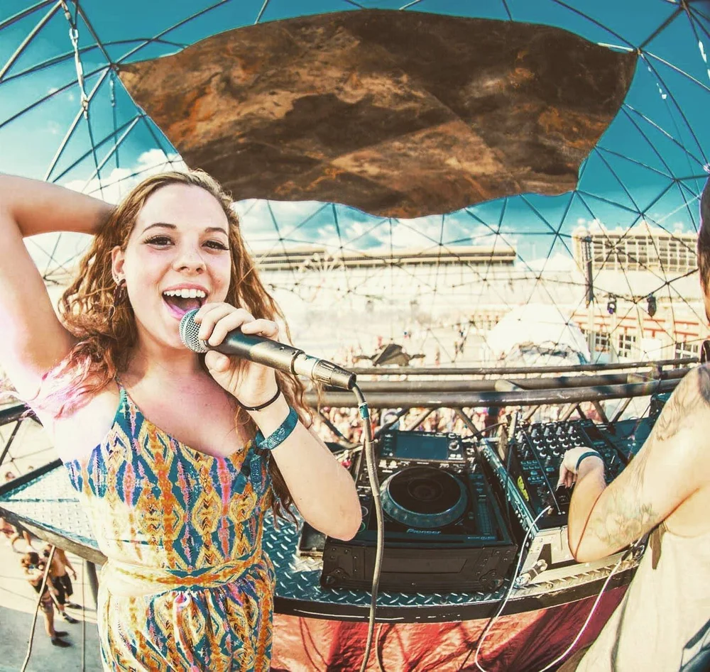 Young woman singing into a microphone on a boat with a geodesic dome overhead, smiling, with a cityscape background.