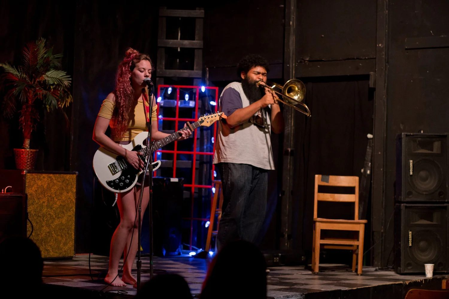 Two musicians perform on stage, one playing guitar and the other playing a trumpet, with stage decorations and equipment visible.