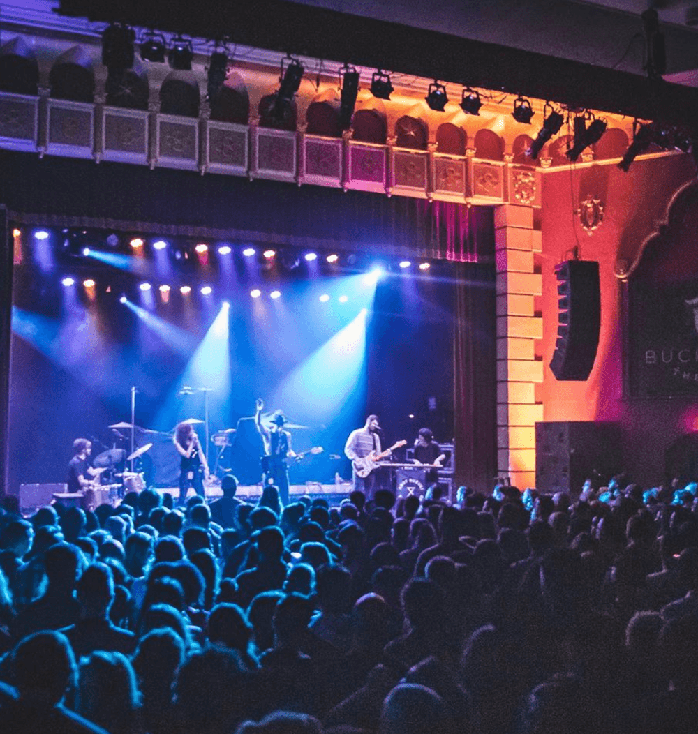 Concert stage with band performing, colorful lighting, audience in foreground, theater interior with ornate architecture, large crowd.