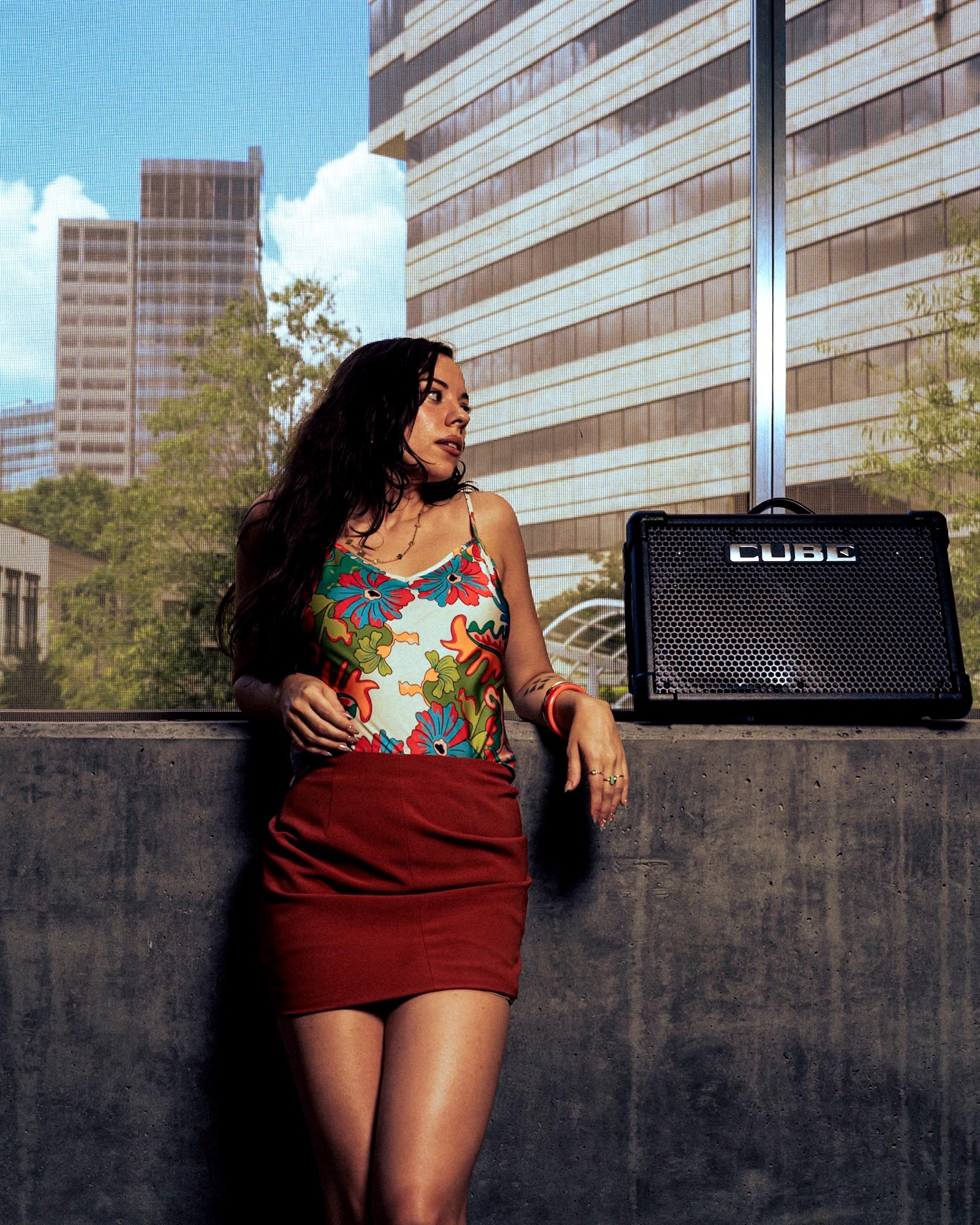 Woman with long dark hair wearing a colorful top and red skirt standing outdoors near a concrete wall with buildings in the background.